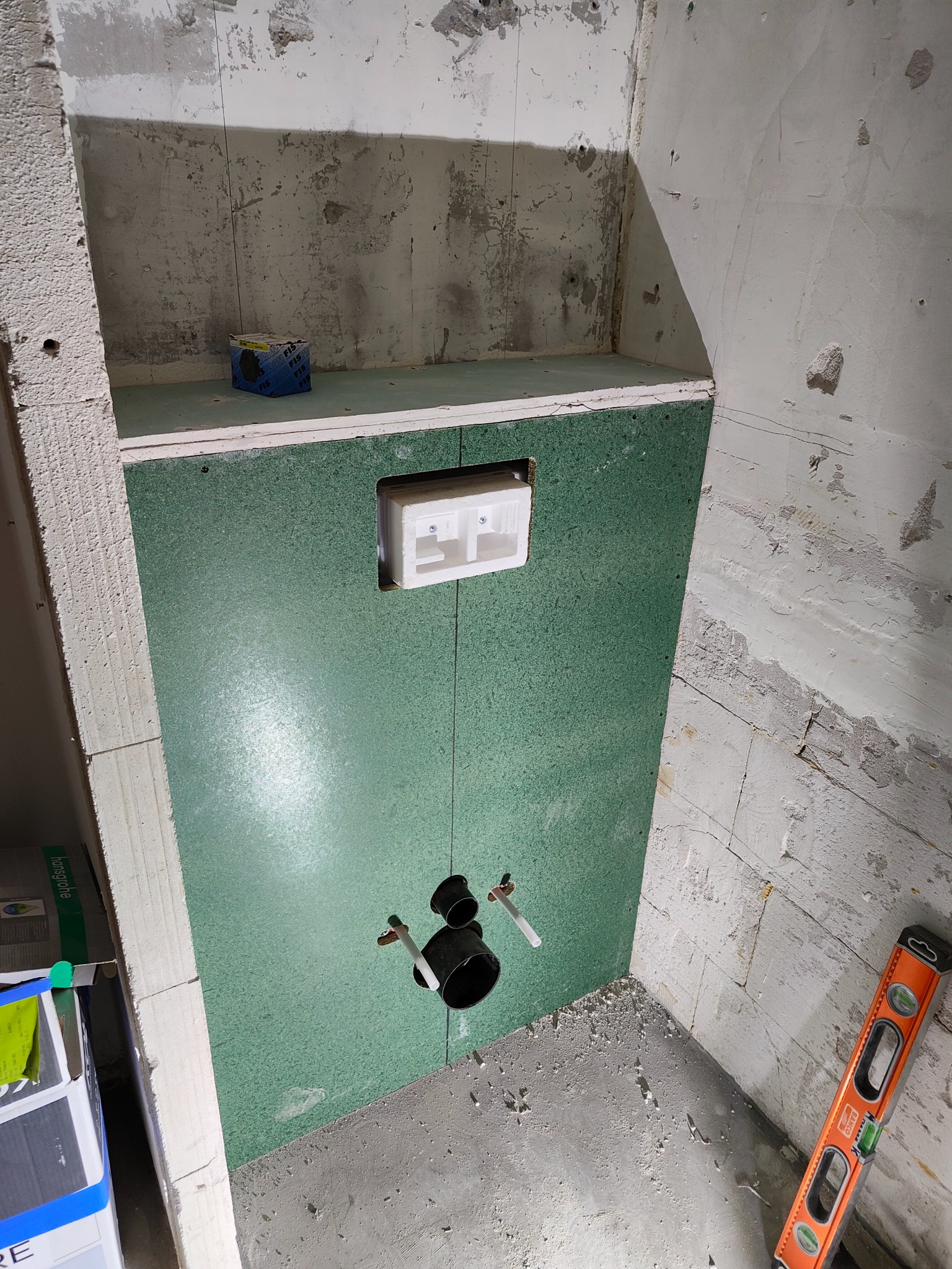 Przygotowanie do montażu stelaża WC na zielonej płycie w surowej łazience. Widoczne rury doprowadzające wodę i poziomica. Etap prac przed wykończeniem.