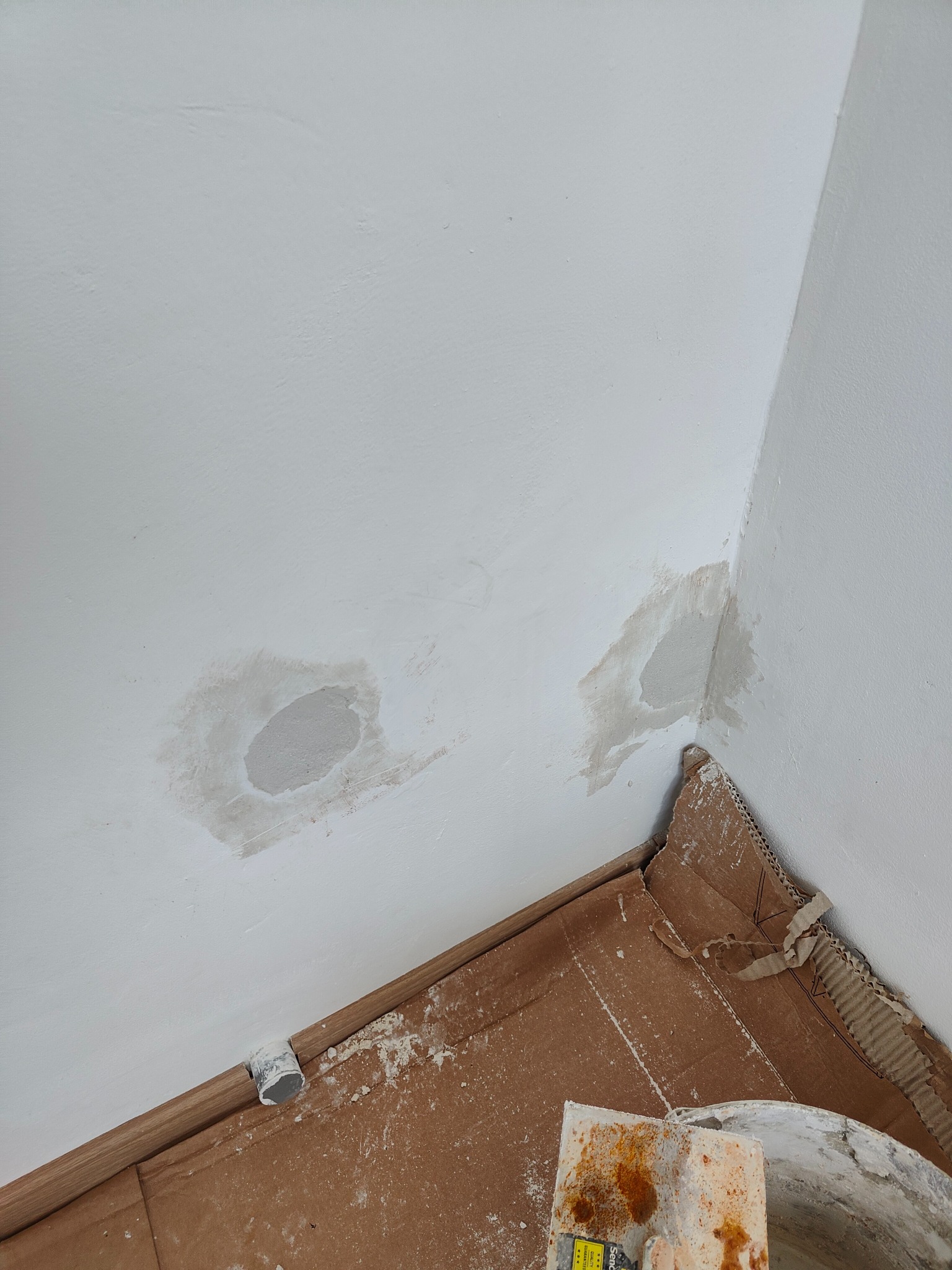 Przygotowanie ściany do malowania: zagruntowane ubytki w rogu pomieszczenia, zabezpieczona podłoga kartonem, widoczne narzędzia malarskie.