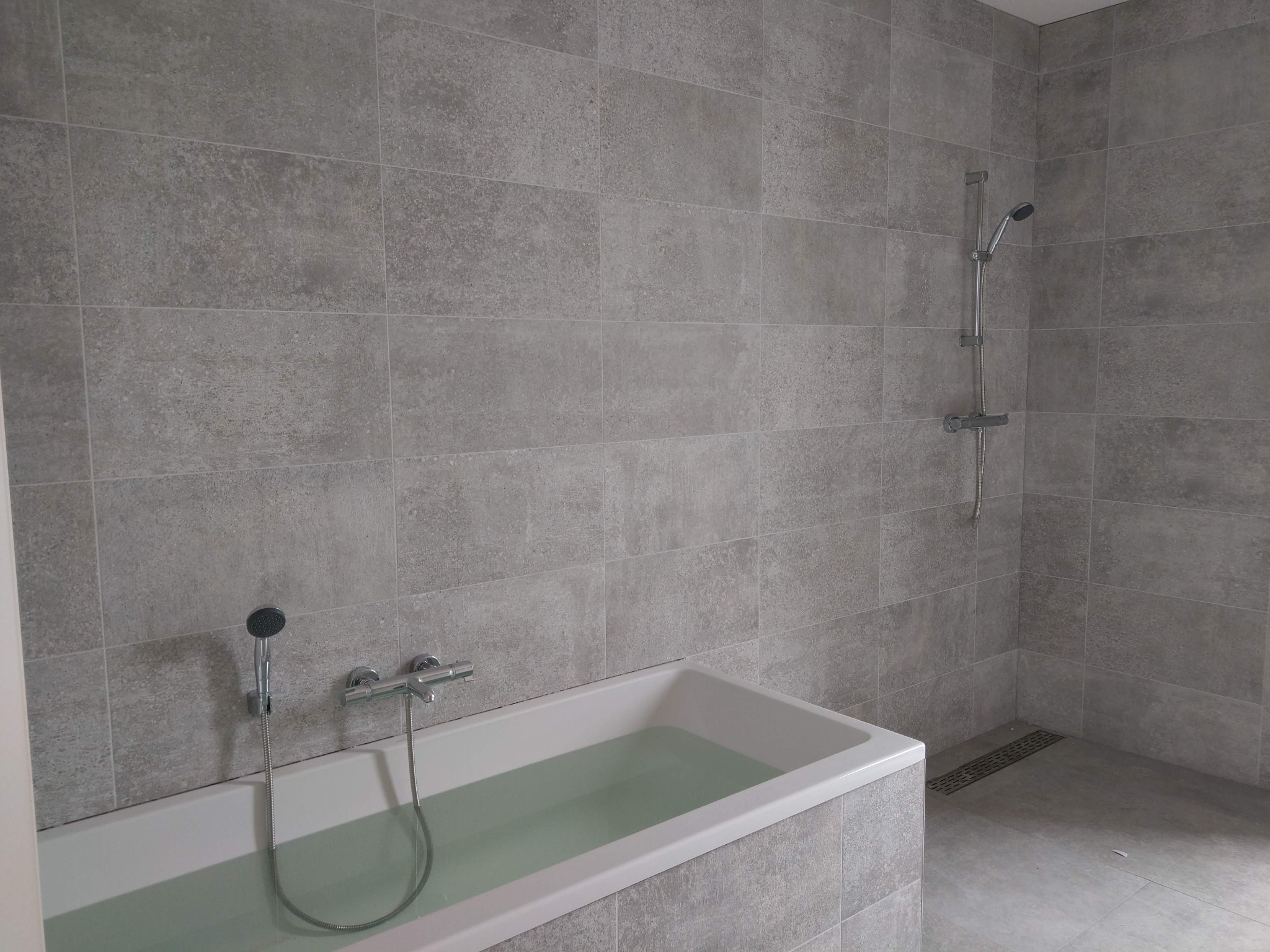 Wykończona łazienka z wanną wypełnioną wodą i szarymi płytkami na ścianach oraz podłodze. Widoczna armatura i odpływ liniowy. Minimalistyczny design.