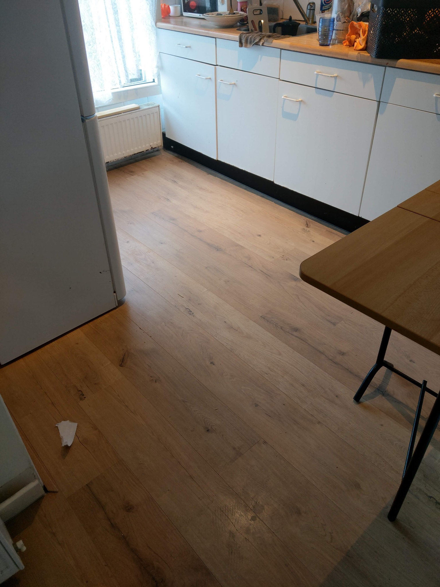 Jasna, drewniana podłoga w kuchni z białymi szafkami i fragmentem stołu. Widoczny fragment lodówki i okna z firanką. Minimalistyczny wystrój, ciepłe oświetlenie.