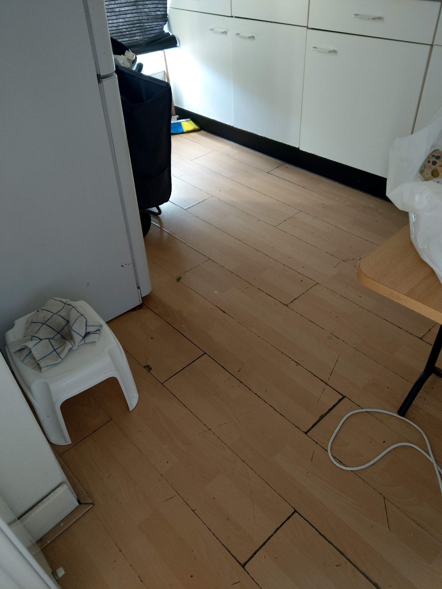 Ukończona podłoga z paneli w kuchni, widoczne szczeliny między panelami, białe szafki w tle, plastikowe krzesło z szarą ścierką i kabel na podłodze.