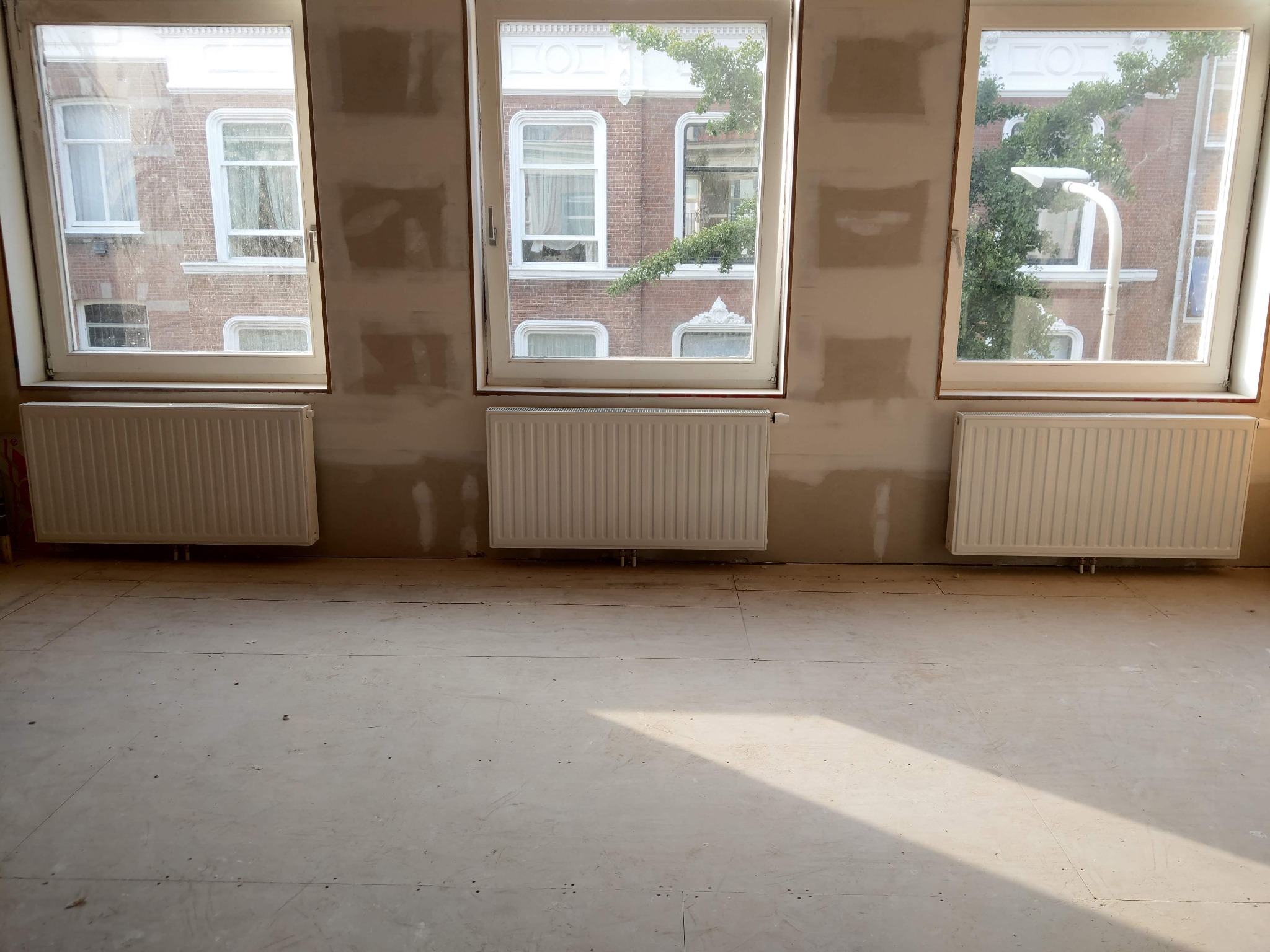 Pokój po remoncie: trzy okna z widokiem na budynek, pod nimi zamontowane grzejniki, ściany przygotowane do malowania, podłoga wyłożona płytami. Jasne wnętrze.