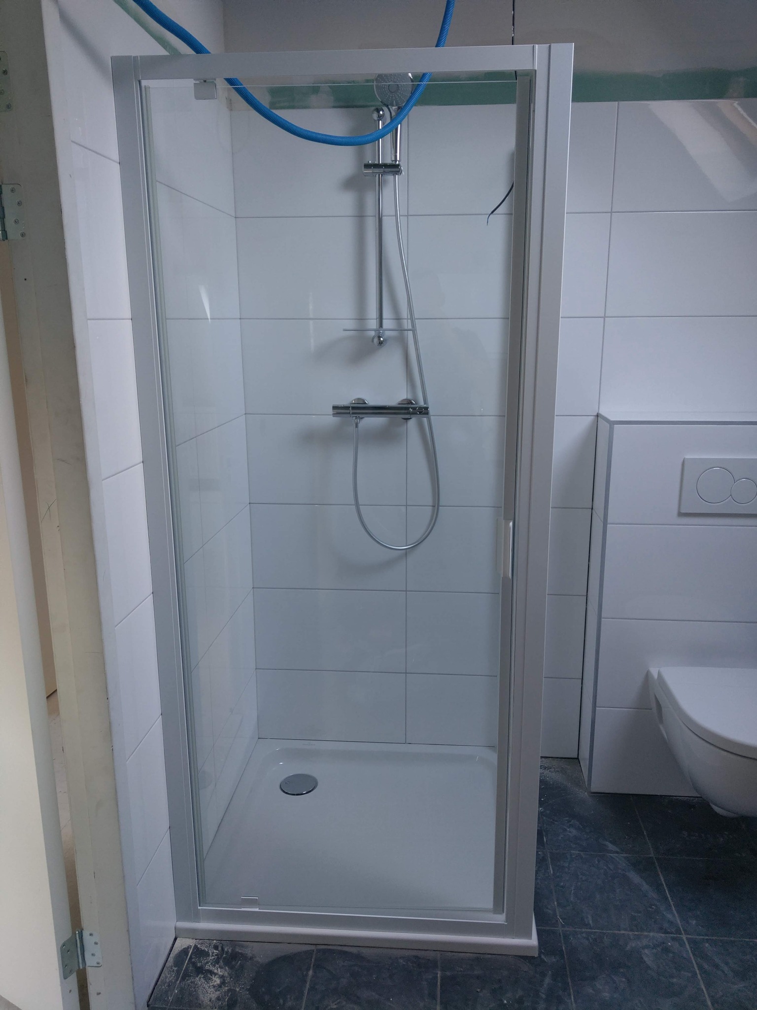 Nowa kabina prysznicowa z brodzikiem i baterią, zamontowana w łazience wyłożonej białymi płytkami. Widoczne niebieskie rury instalacji wodnej.