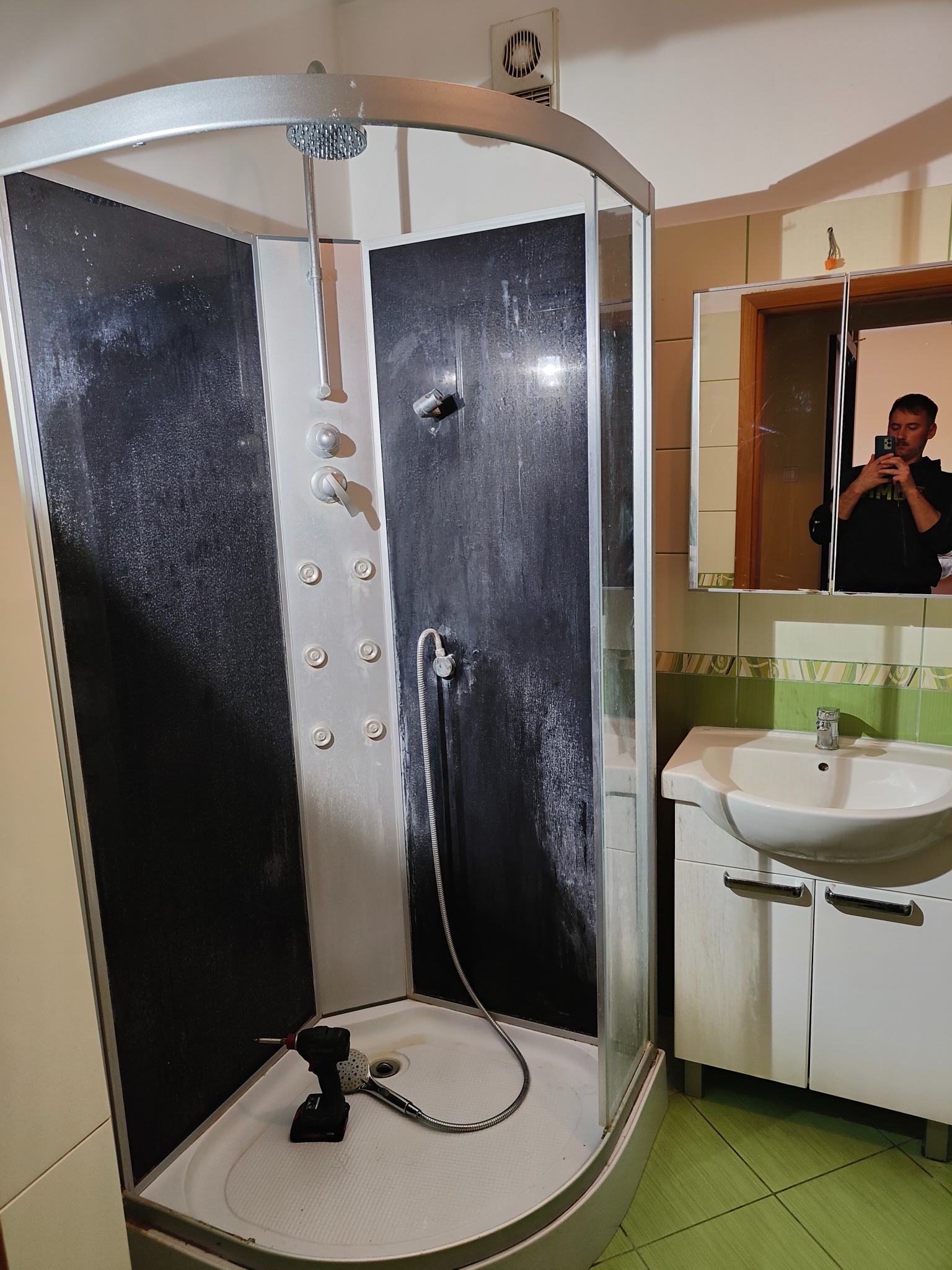 Kabina prysznicowa z hydromasażem w trakcie montażu. W brodziku wiertarko-wkrętarka. W lustrze odbija się osoba robiąca zdjęcie. Zielona podłoga.