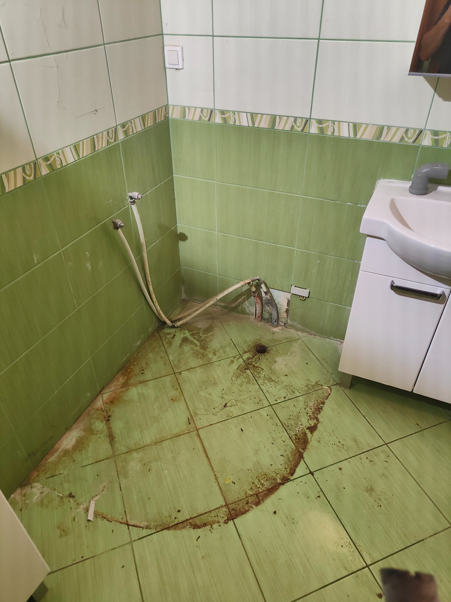 Łazienka w trakcie remontu: zielone płytki, biała szafka pod umywalką, wystające rury hydrauliczne. Widoczne ślady po demontażu i zabrudzenia na podłodze.