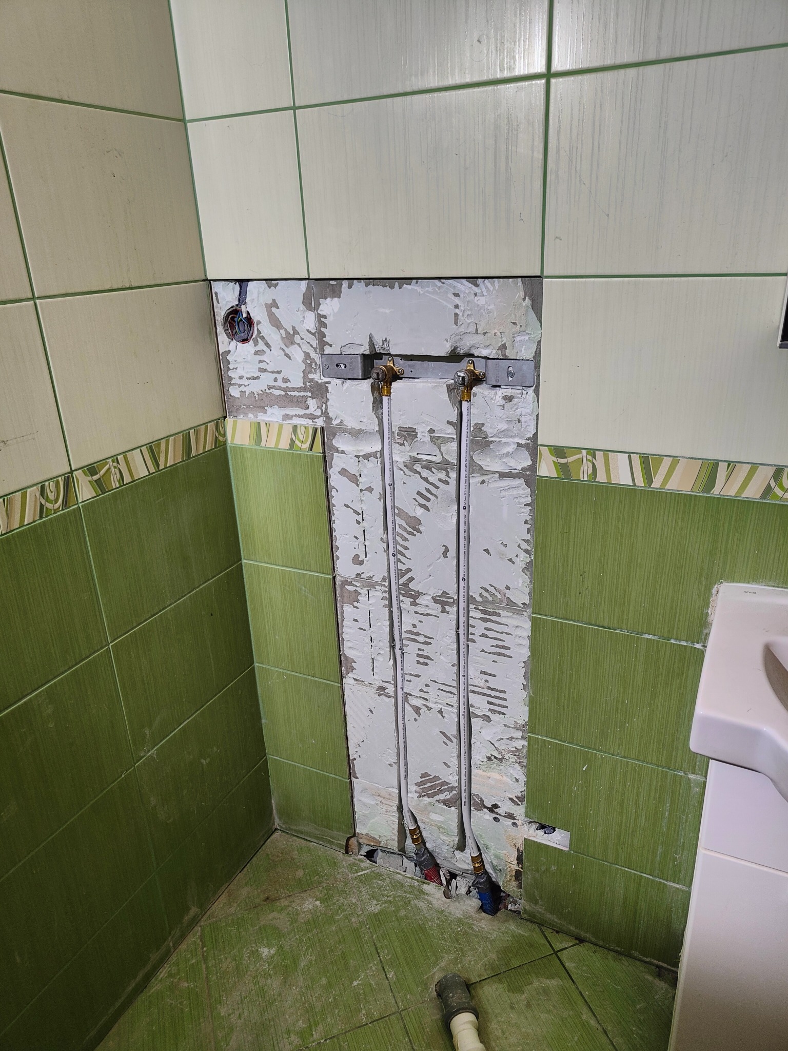 Fragment łazienki w trakcie remontu. Widoczne rury instalacji wodnej przymocowane do ściany, częściowo bez płytek, z zielonymi płytkami w tle i fragmentem umywalki.