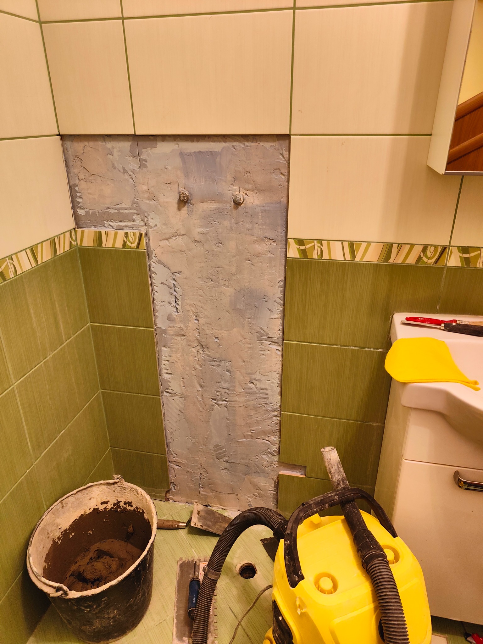 Fragment ściany w łazience z usuniętymi płytkami, przygotowany do dalszych prac. Widoczne narzędzia i wiadro z zaprawą. Prace remontowe w toku.
