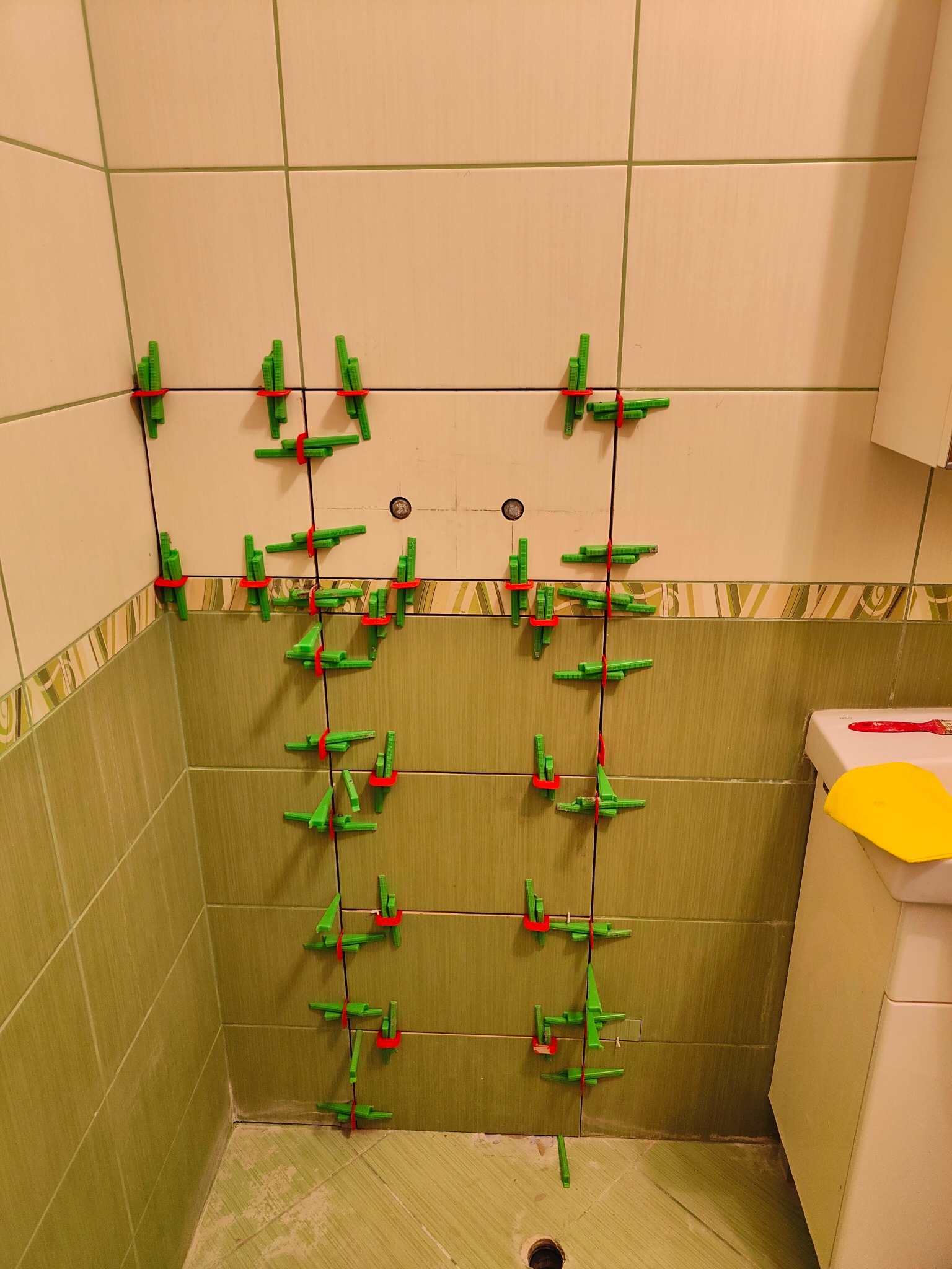 Ściana w łazience z zielonymi i beżowymi płytkami, na których zamontowano system poziomowania płytek z zielonymi klipsami i czerwonymi klinami. Widoczny fragment toalety i narzędzia.