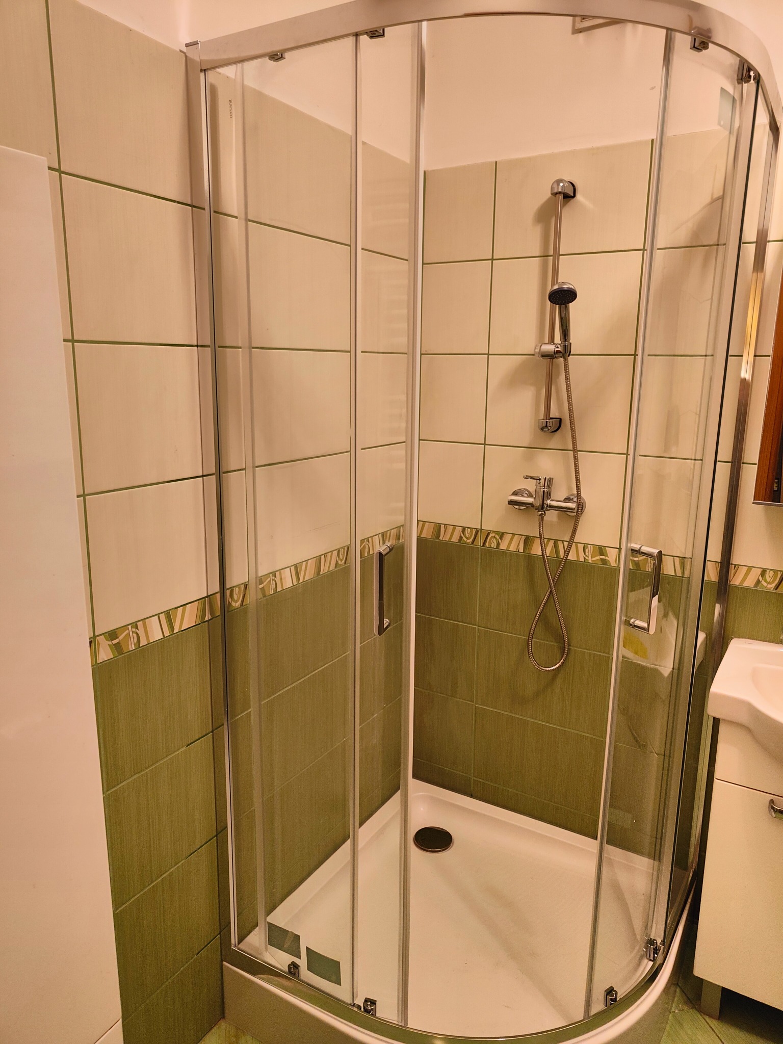 Kabina prysznicowa narożna z chromowanymi profilami i transparentnym szkłem, widoczna armatura i zielono-beżowe płytki w łazience. Umywalka z szafką po prawej.