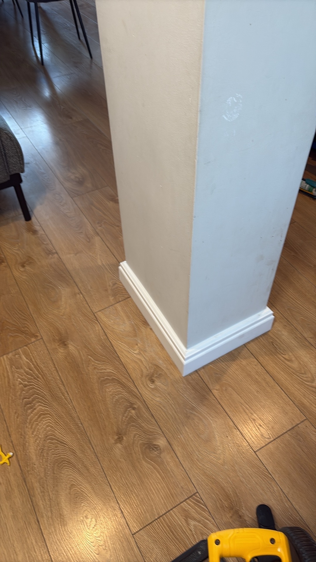 Biała kolumna z dekoracyjną listwą przypodłogową na drewnianej podłodze. Fragment narzędzia w rogu kadru. Widoczne ślady po remoncie na kolumnie.