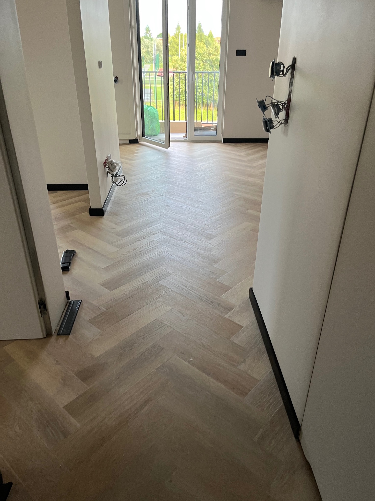 Jasne panele ułożone w jodełkę francuską w nowym mieszkaniu z otwartym balkonem. Widoczne niedokończone instalacje elektryczne i listwy przypodłogowe.