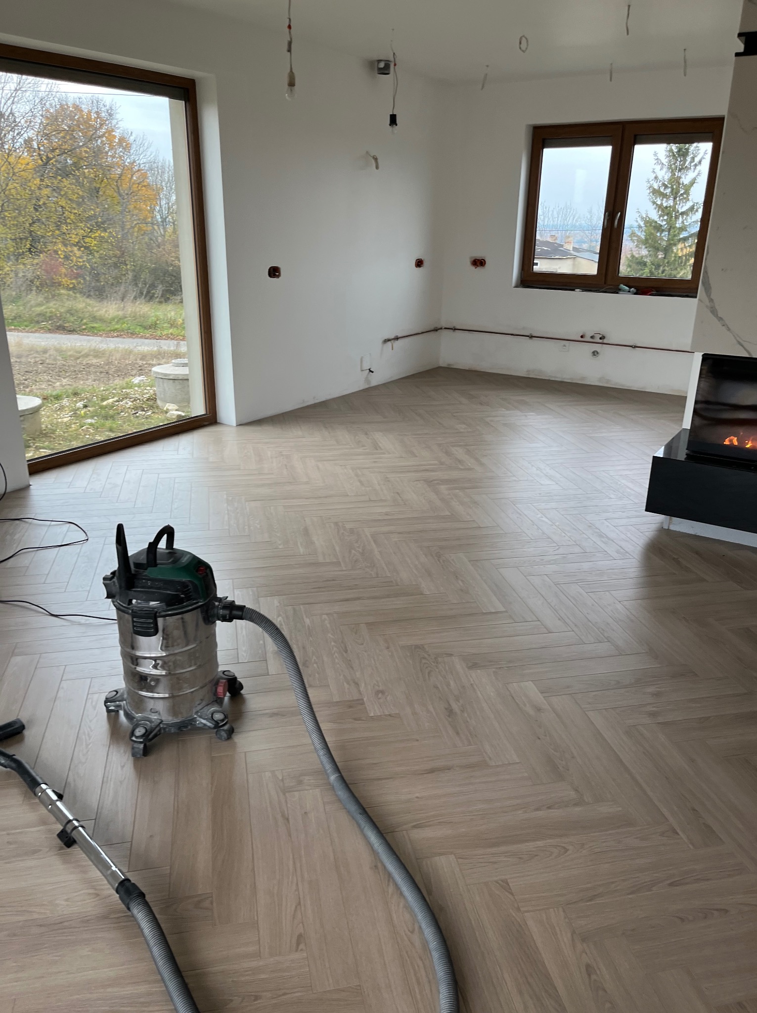 Salon po ułożeniu parkietu w jodełkę, odkurzacz przemysłowy na podłodze, duże okna z widokiem na zewnątrz, minimalistyczny kominek. Świeżo wykończone wnętrze.