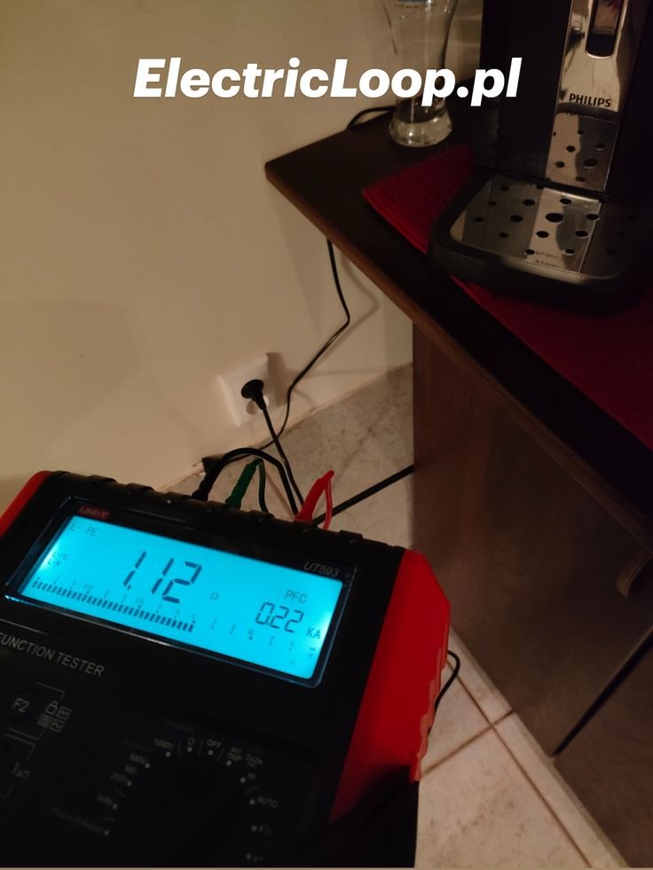 Miernik elektryczny UT593 z podłączonymi przewodami do gniazdka elektrycznego, wskazujący wartości pomiarowe na wyświetlaczu. Obok ekspres do kawy.