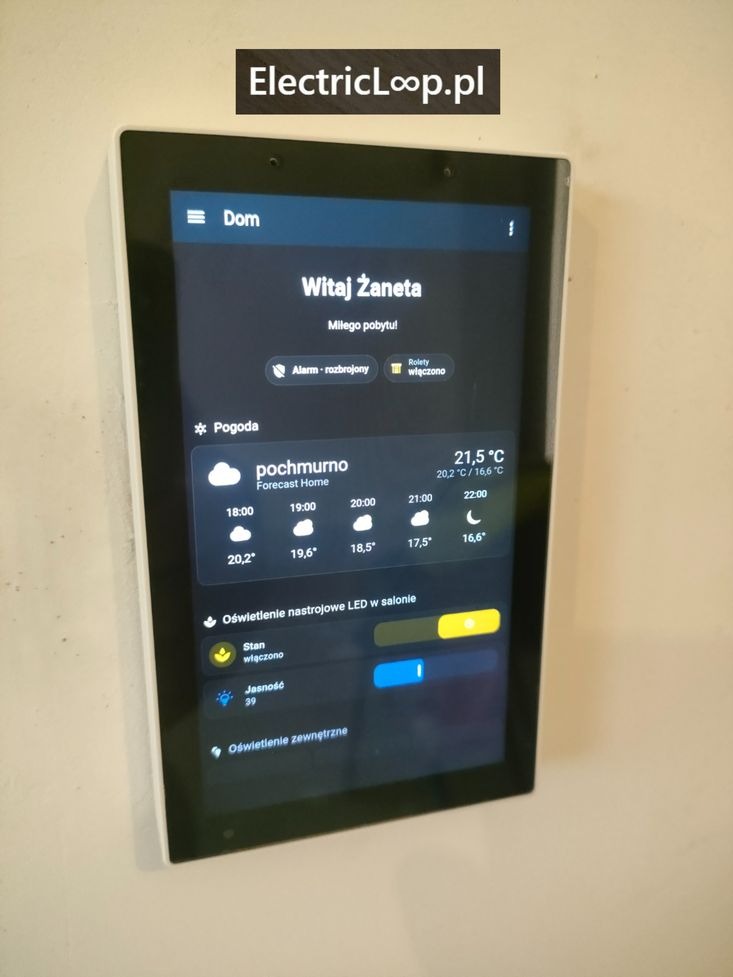 Panel dotykowy na ścianie prezentujący inteligentny system zarządzania domem, z widocznymi informacjami o pogodzie i sterowaniu oświetleniem.