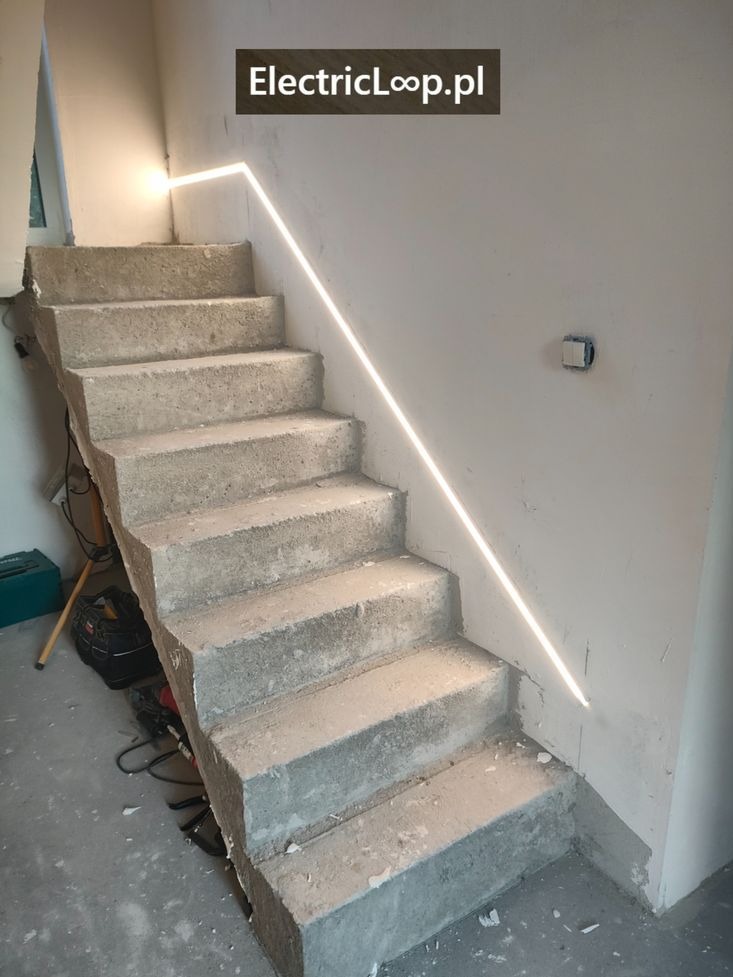 Surowe betonowe schody z subtelnym oświetleniem LED wzdłuż ściany, podkreślającym minimalistyczny design wnętrza w trakcie wykończenia.