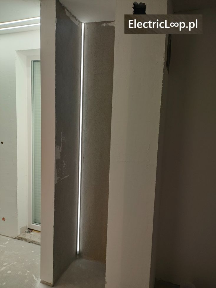 Wnęka z oświetleniem LED w ścianie, widoczne niedokończone wykończenie, surowy beton i fragment okna. Minimalistyczny design z subtelnym akcentem świetlnym.