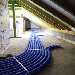 HydroProjekt Białystok Mateusz Banel - Niebieskie rury wentylacyjne ułożone w rzędzie na poddaszu, widoczne drewniane elementy konstrukcji dachu i izolacja termiczna. Instalacja rekuperacji w trakcie montażu.