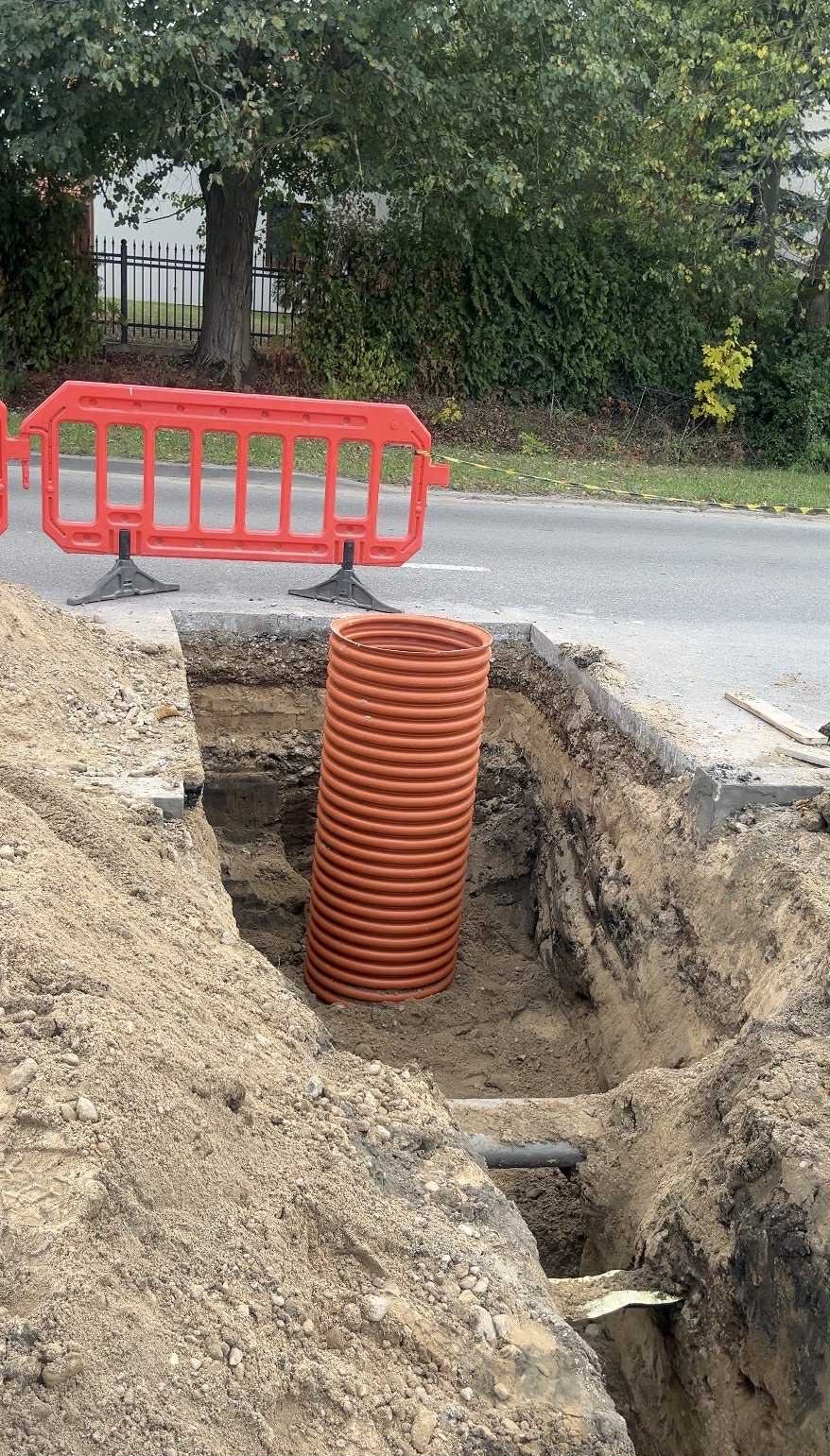 Pomarańczowa rura karbowana w wykopie ziemnym, zabezpieczona barierą drogową. Widoczne warstwy gruntu i fragmenty instalacji. Prace ziemne przy drodze.