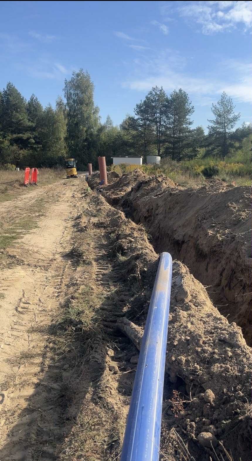 Wykop pod instalację z widocznymi rurami, pomarańczowymi słupkami i koparką w tle, na tle drzew i błękitnego nieba. Prace ziemne w toku.