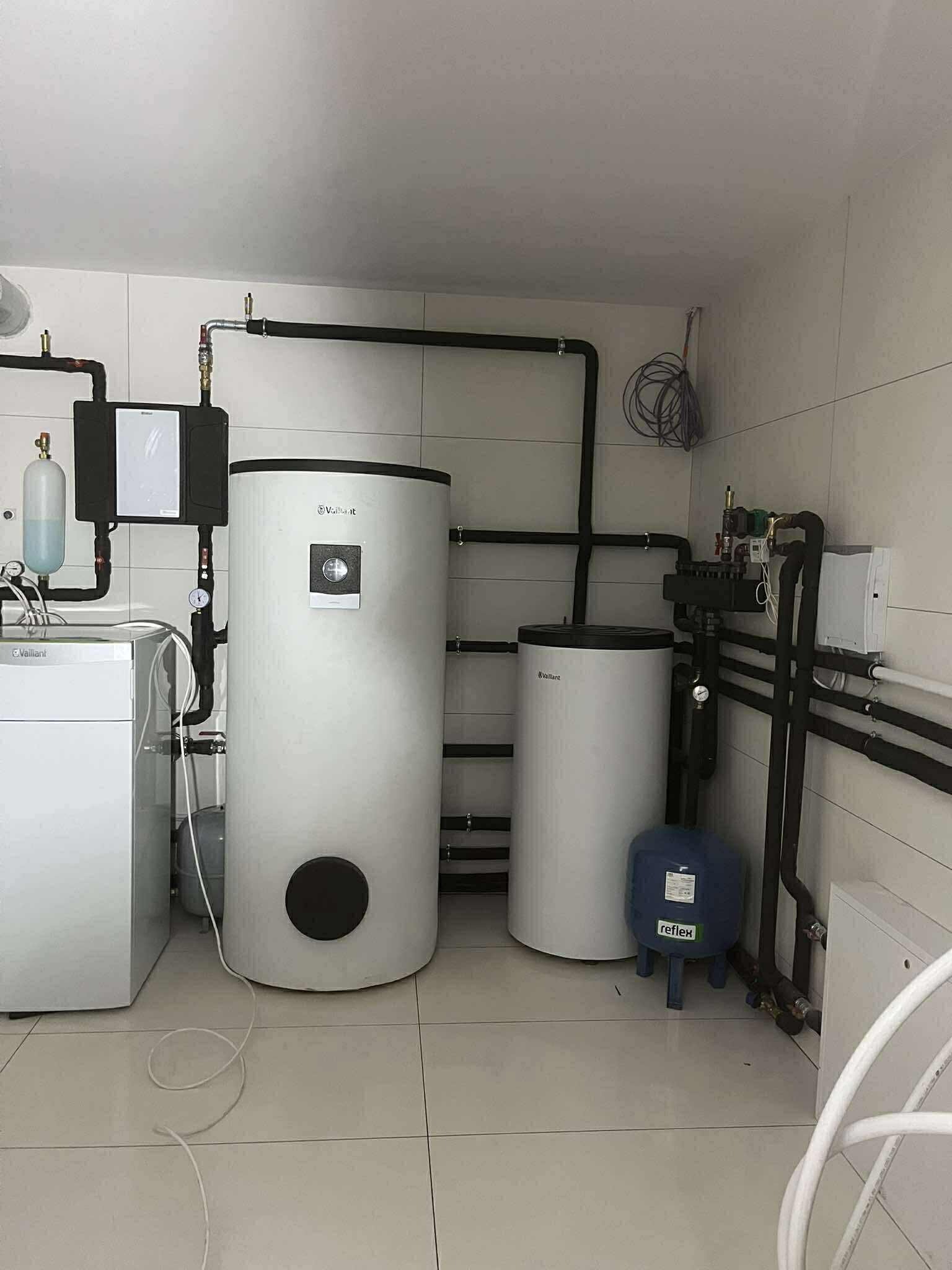 Białe zbiorniki Vaillant w kotłowni z czarnymi rurami, manometrami i niebieskim zbiornikiem Reflex. Ściany wyłożone jasnymi płytkami, przewody zwisają z sufitu.