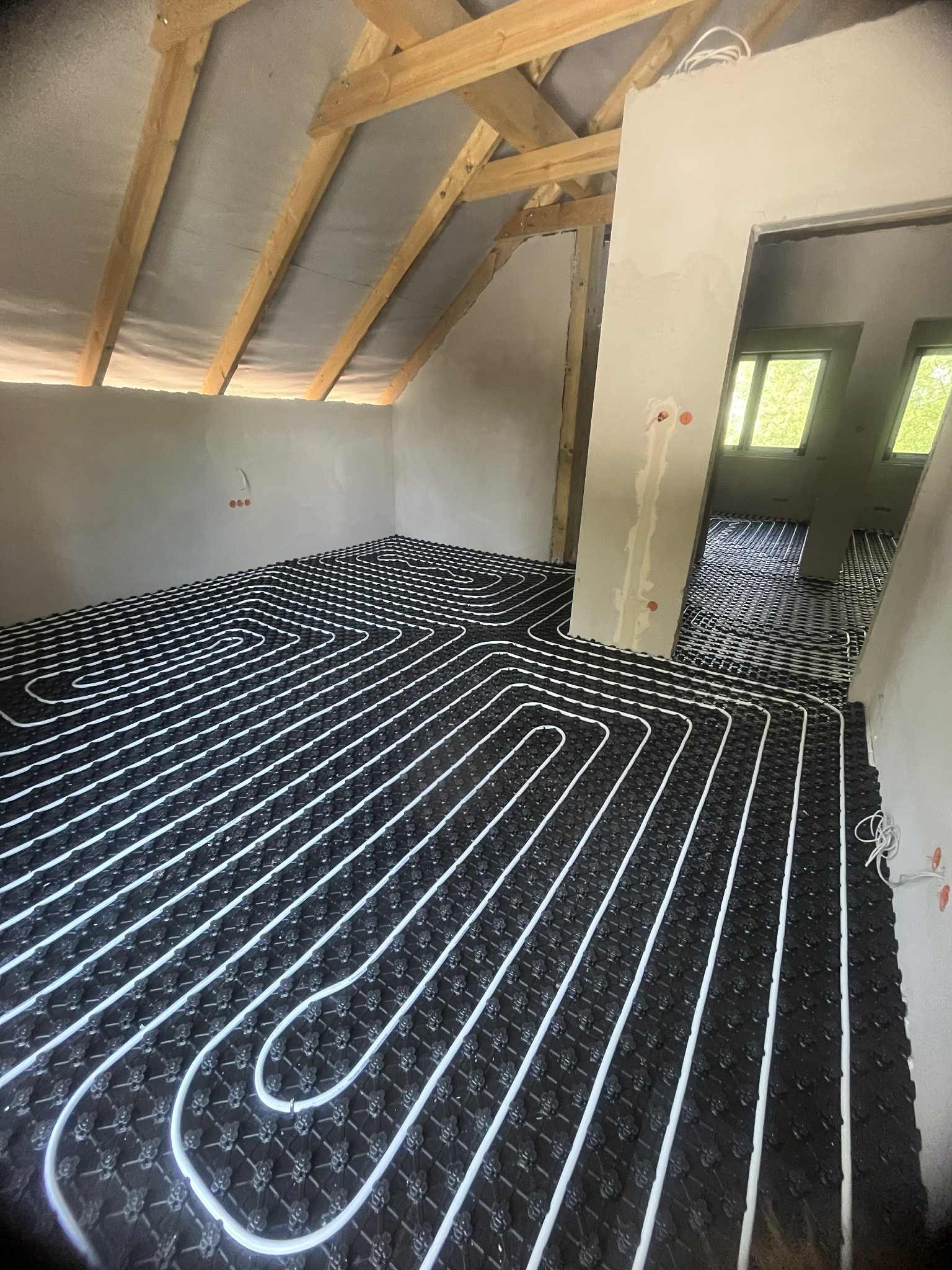 Podłogówka w poddaszu: białe rury grzewcze ułożone na czarnej macie izolacyjnej, widoczne krokwie dachowe i surowy tynk. Instalacja przed wylaniem posadzki.