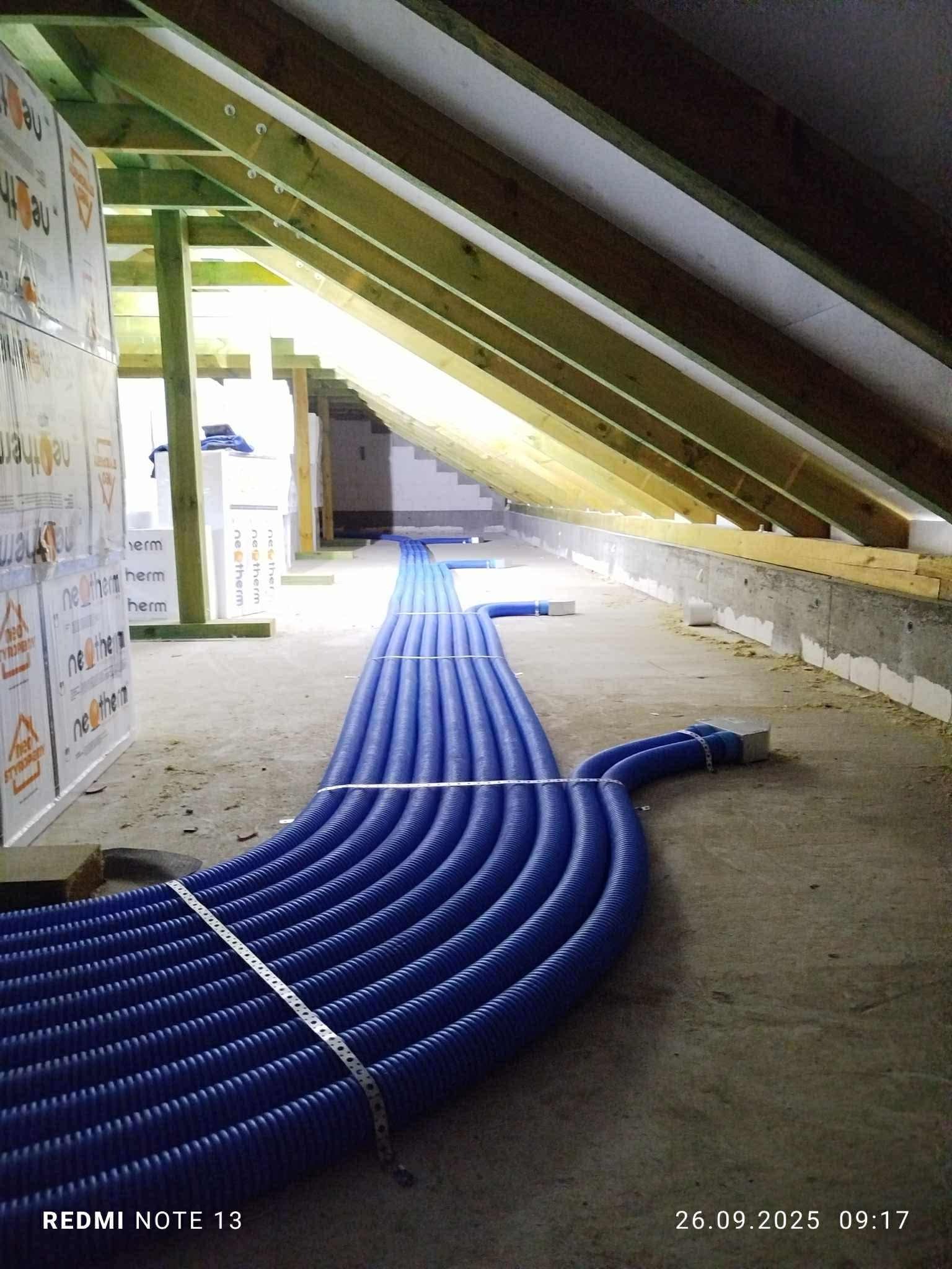 Niebieskie rury wentylacyjne ułożone w rzędzie na poddaszu, widoczne drewniane elementy konstrukcji dachu i izolacja termiczna. Instalacja rekuperacji w trakcie montażu.