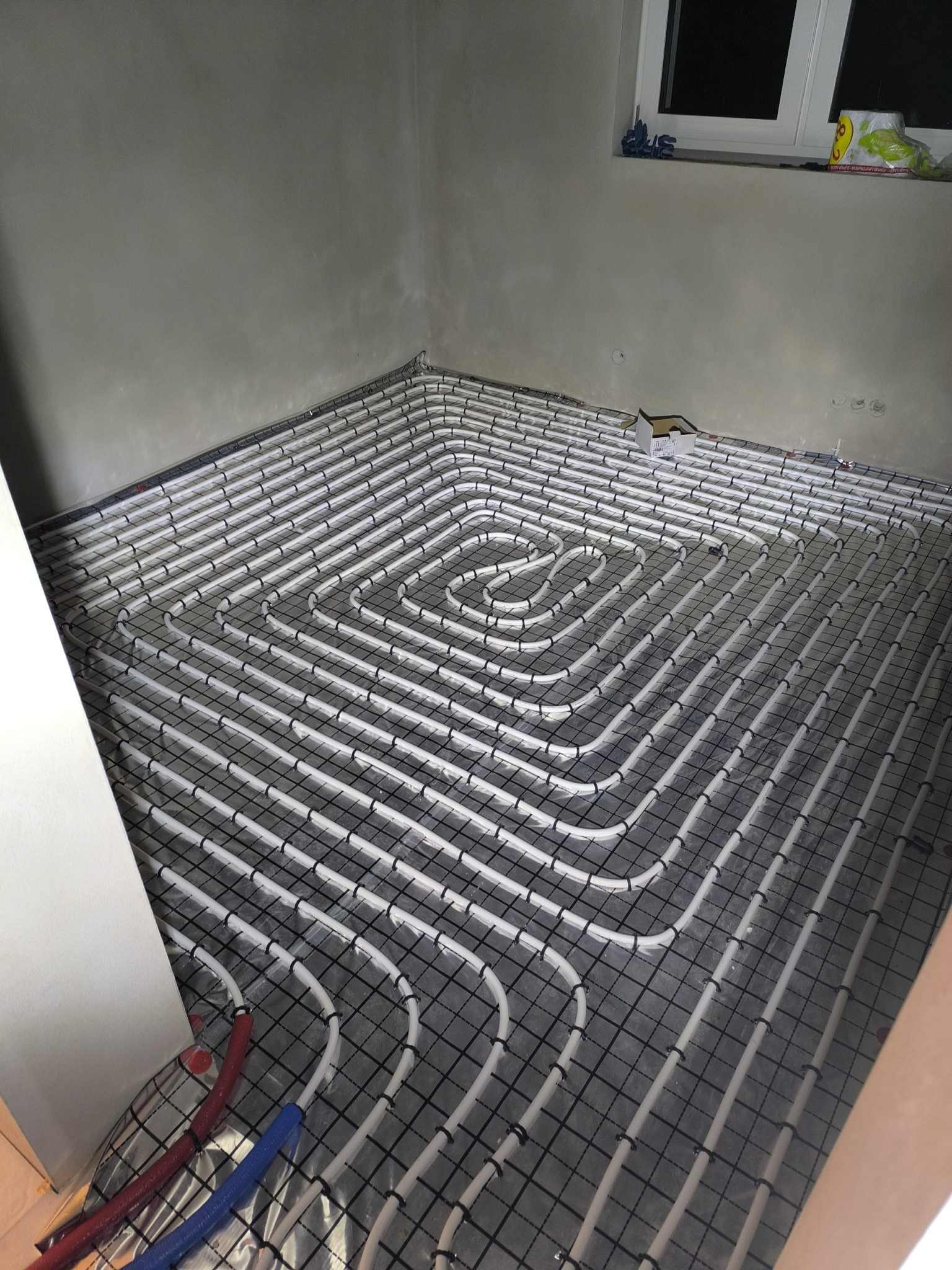 Ułożona instalacja ogrzewania podłogowego w kształcie spirali na czarnej siatce, widoczne rury doprowadzające w kolorze niebieskim i czerwonym, surowy stan ścian.