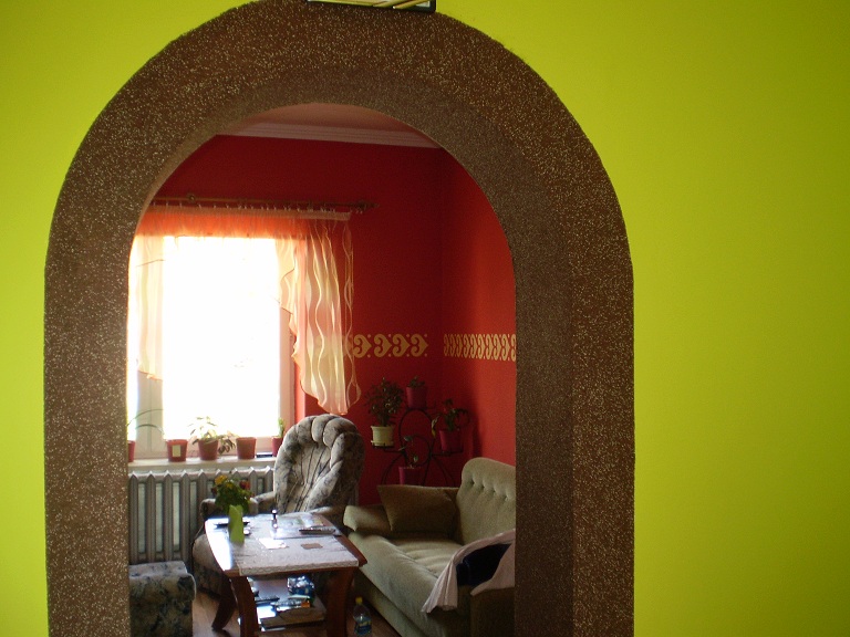 Arched doorway with textured finish, leading to a living room with red walls and patterned curtains, viewed from a room with lime green walls.