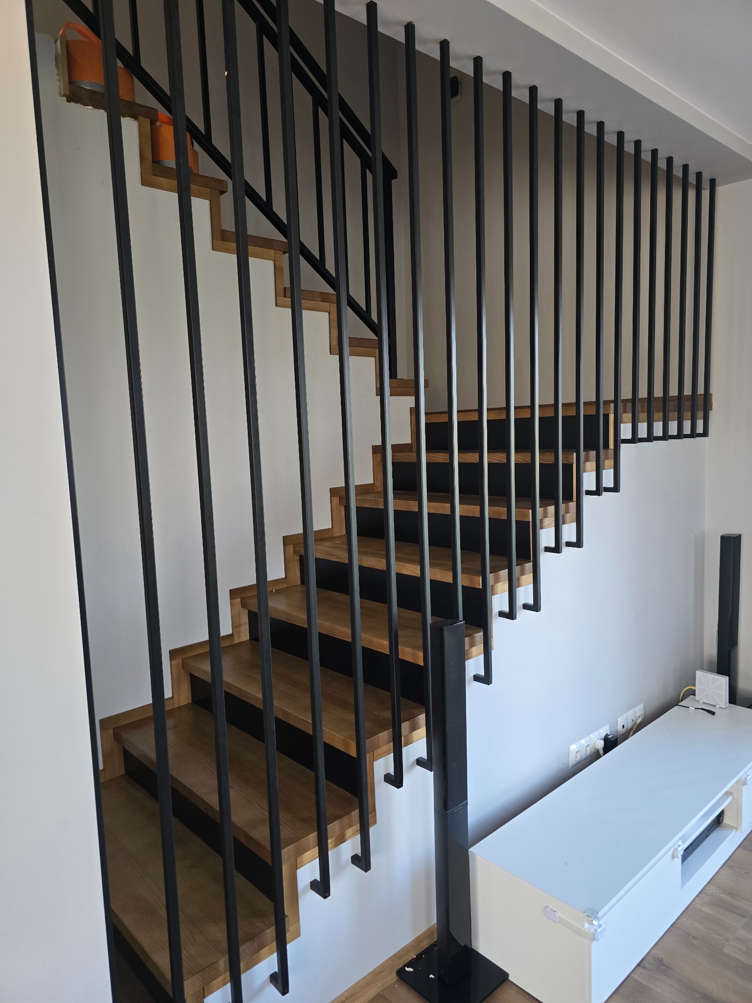 Nowoczesne schody z drewnianymi stopniami i czarną, minimalistyczną balustradą z pionowych prętów. Widok z dołu, w tle biała ściana i meble.