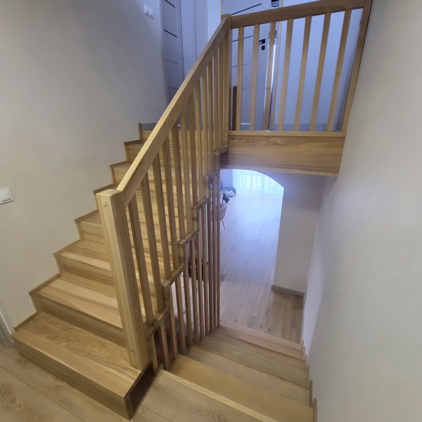 Jasne, drewniane schody z balustradą w nowoczesnym domu. Widok z góry na dół, z delikatnym oświetleniem w tle. Minimalistyczny design i naturalne wykończenie drewna.