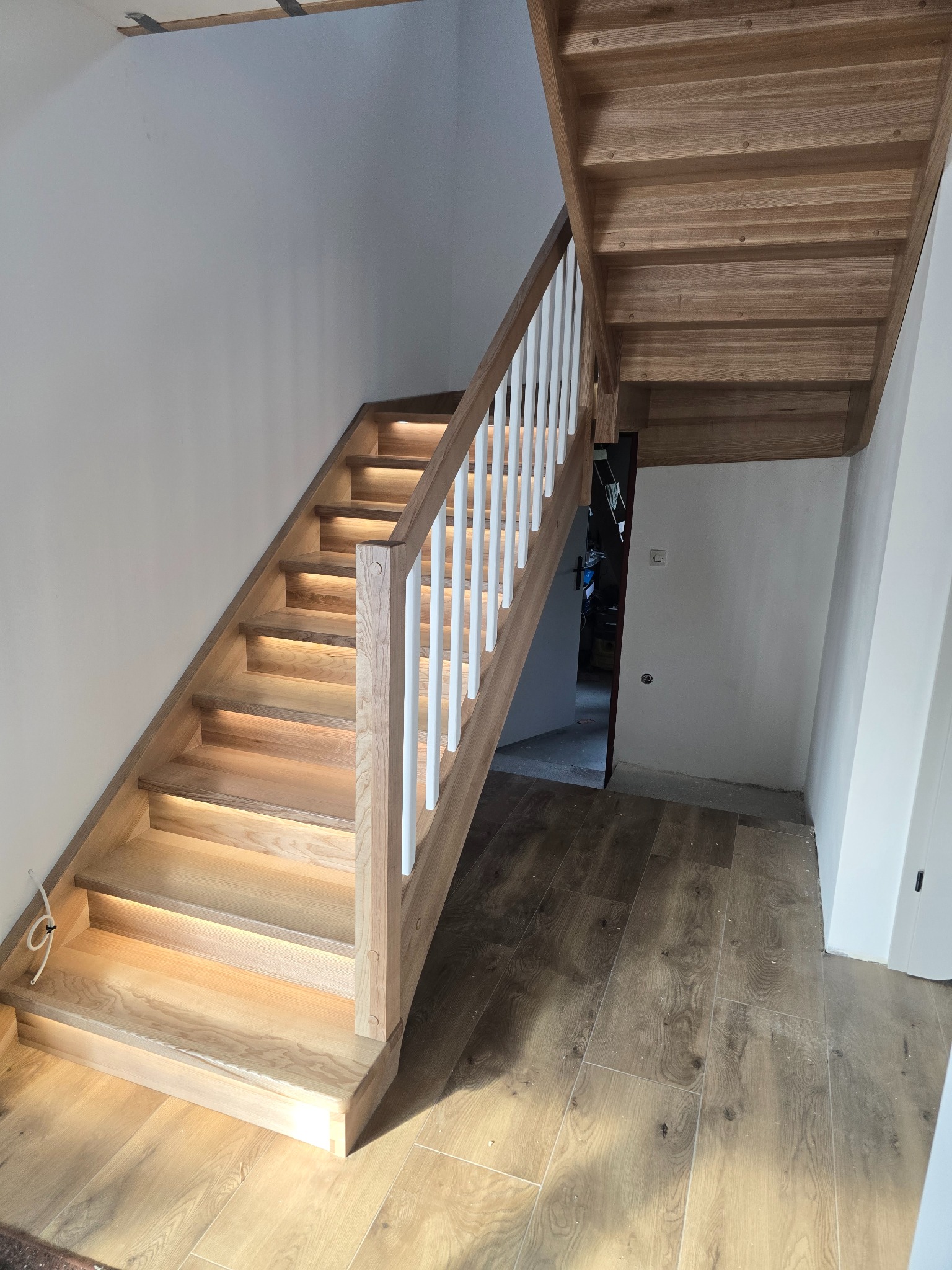 Jasne, drewniane schody z białymi balustradami i subtelnym podświetleniem LED, prowadzące na górę. Widoczne drewniane wykończenie i parkiet na podłodze.