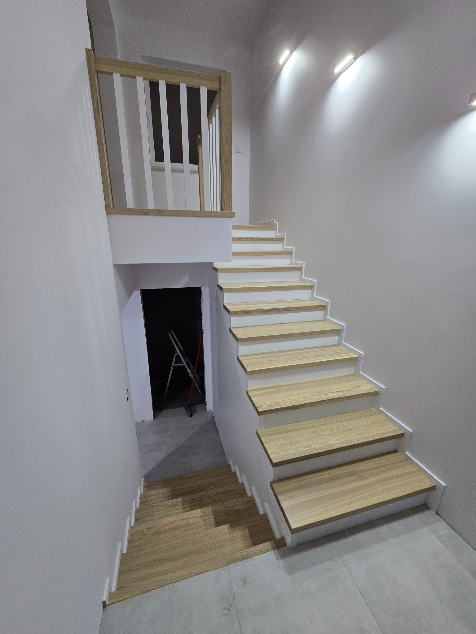 Jasne, nowoczesne schody z drewnianymi stopniami i białą balustradą, prowadzące w dół. W tle widoczna drabina w ciemnym pomieszczeniu. Minimalistyczny design.