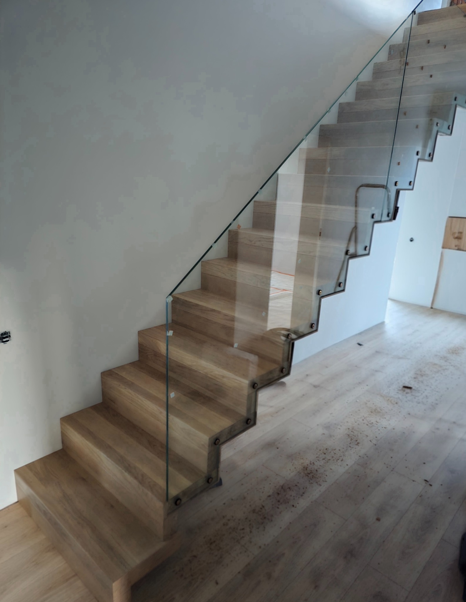 Nowoczesne schody z jasnego drewna z szklaną balustradą, mocowaną na metalowych elementach. Widok z dołu, na podłodze drobne wióry.