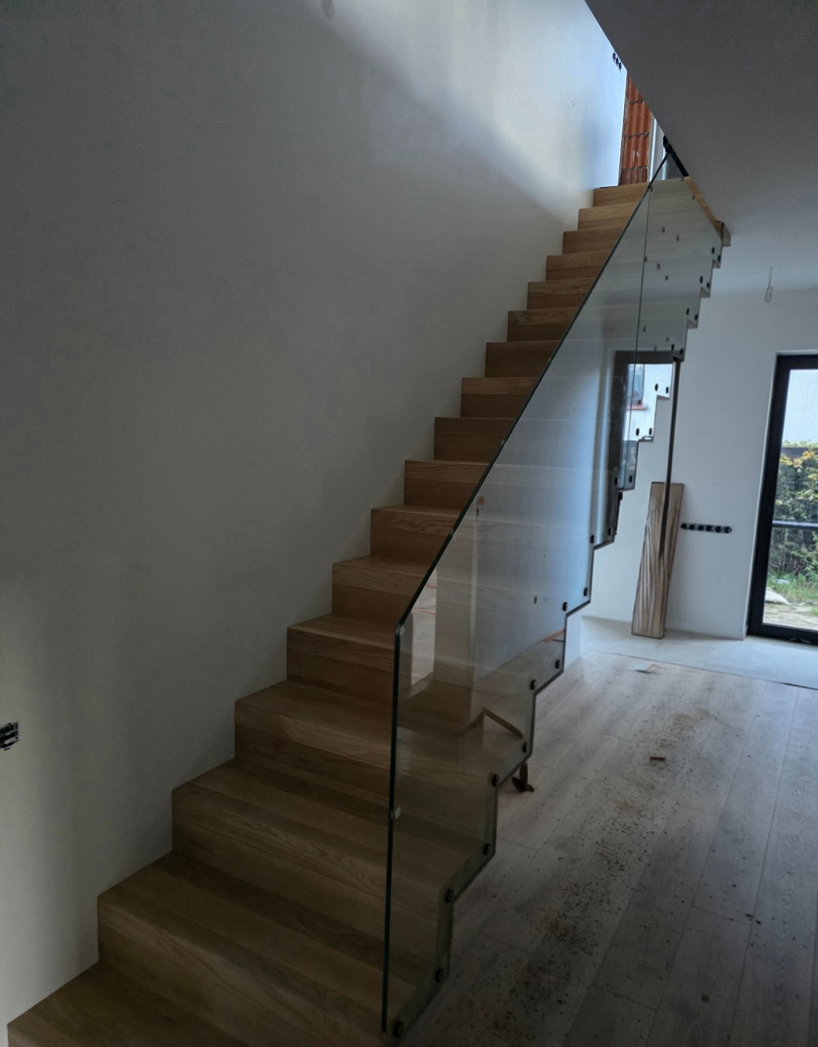 Drewniane schody z nowoczesną szklaną balustradą, widok z dołu. Naturalne oświetlenie podkreśla strukturę drewna i minimalistyczny design.