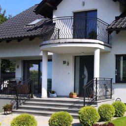 HAPIO GRZEGORZ KOCZERBA - Elegancki dom z balkonem i czarną stolarką okienną, widok z zewnątrz. Schody wejściowe prowadzą do drzwi z numerem 10, otoczone zielenią.