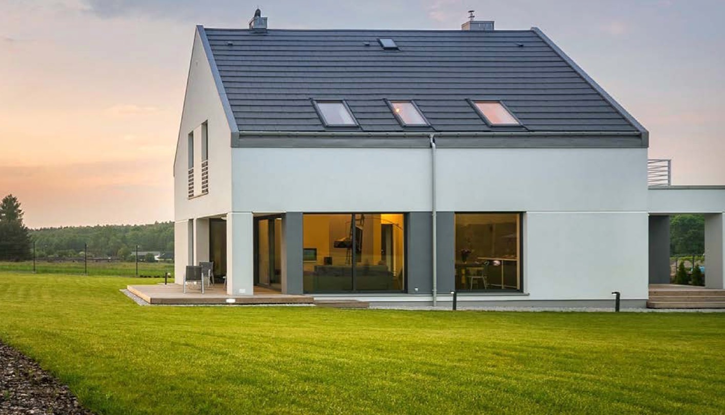 Nowoczesny dom z dużymi oknami i przesuwnymi drzwiami tarasowymi, otoczony zielonym trawnikiem, uchwycony w ciepłym świetle zachodzącego słońca.