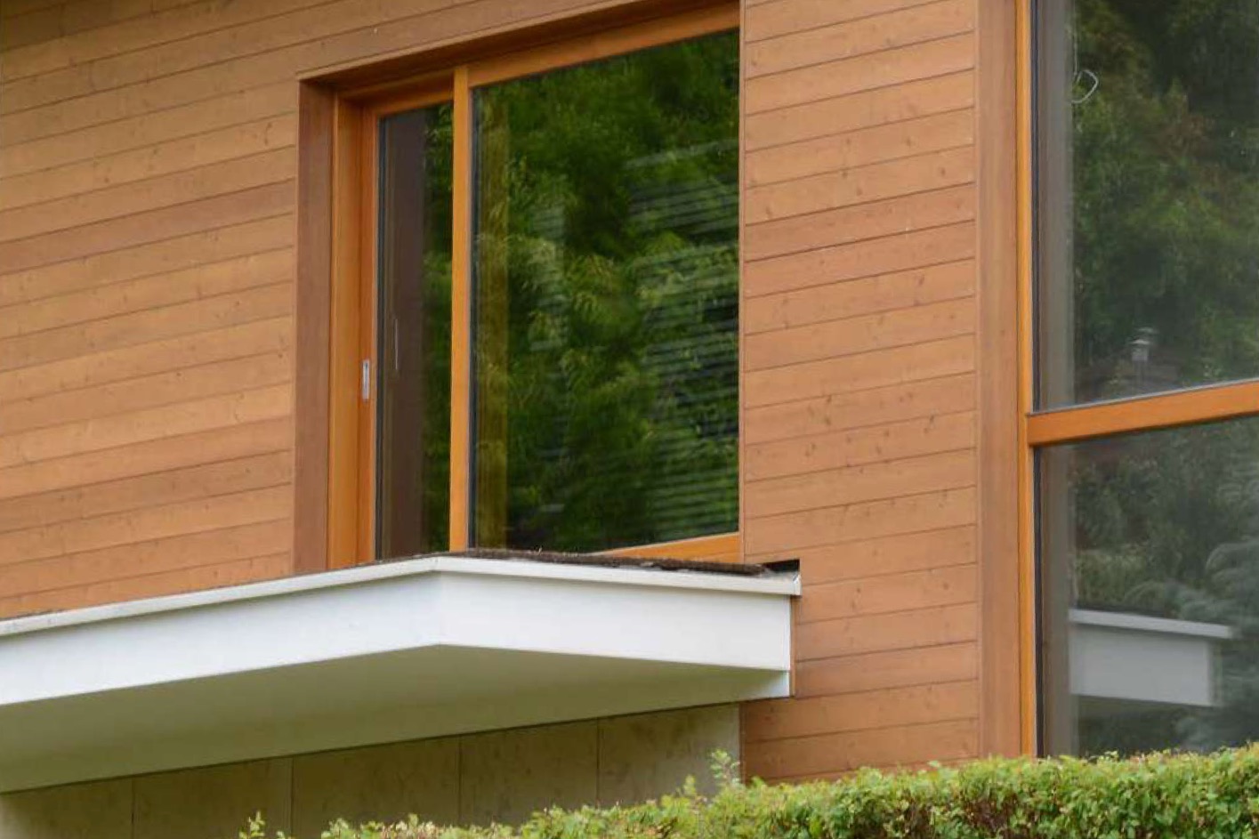 Drewniana elewacja budynku z dużym oknem przesuwnym i balkonem. Widok na zieleń za oknem. Naturalne światło, ciepłe odcienie drewna.