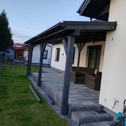 Usługi dekarskie Karol Próchniewicz - Zadaszony taras z ciemnymi elementami drewnianymi i szarą kostką brukową. Widoczne rury spustowe, meble ogrodowe i trawnik. Ujęcie z boku, w tle zabudowa jednorodzinna.