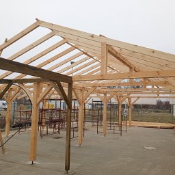 Usługi dekarskie Karol Próchniewicz - Surowa drewniana konstrukcja dachu z widoczną więźbą, słupami i belkami, na tle szarego nieba. Elementy metalowe w tle. Widok z poziomu gruntu.