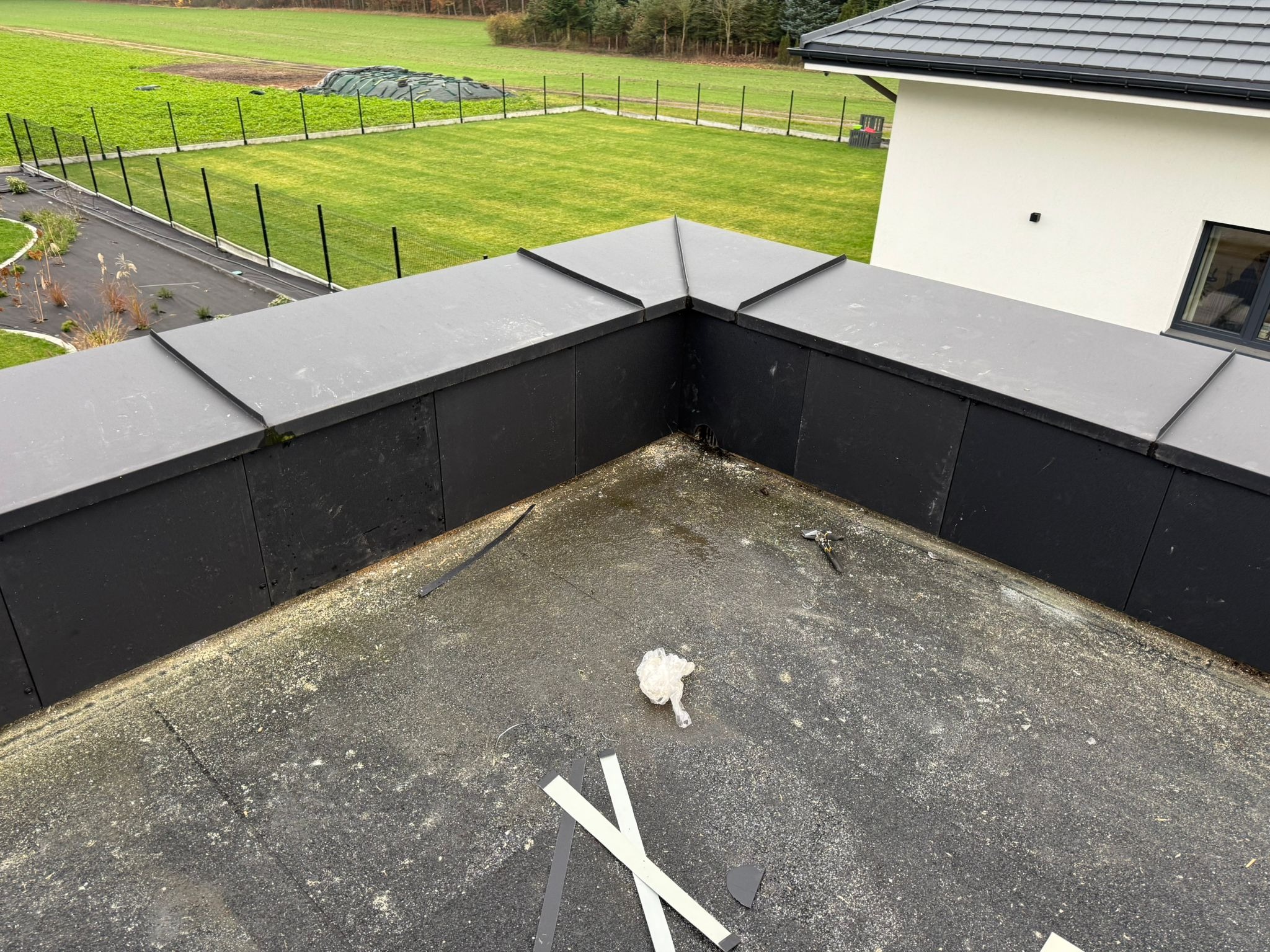 Czarny, płaski dach z obróbkami blacharskimi na tle zielonego trawnika i białego budynku. Widoczne elementy wykończeniowe i resztki materiałów po pracy.