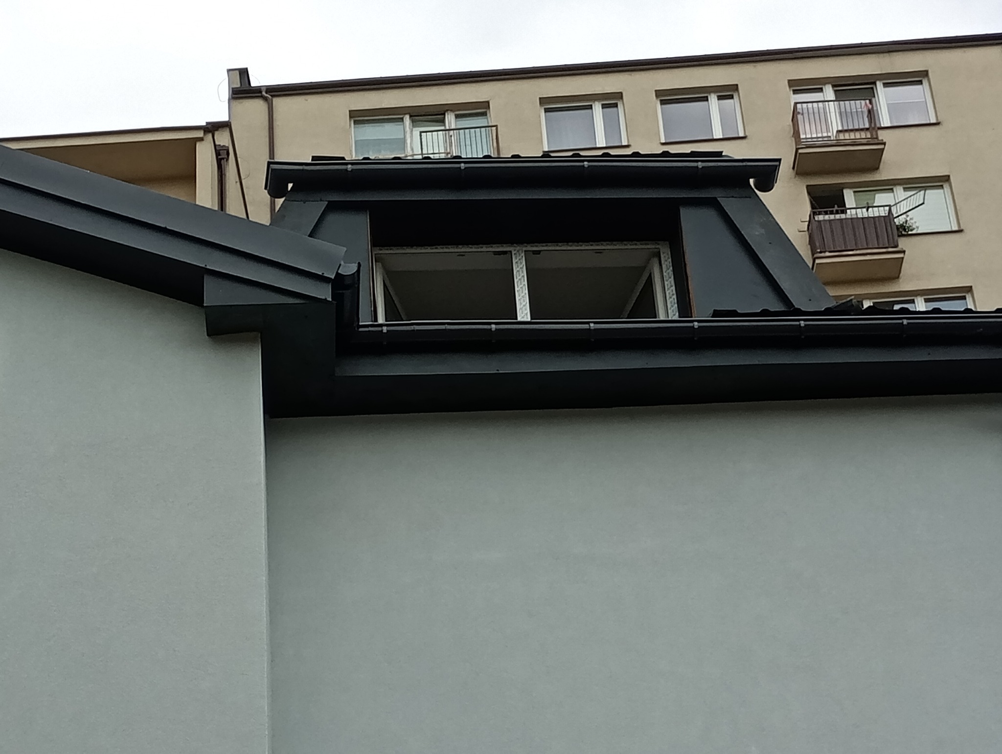 Czarny dach z oknem dachowym w trakcie montażu, widok z dołu na tle budynku mieszkalnego. Rynna wzdłuż krawędzi dachu.