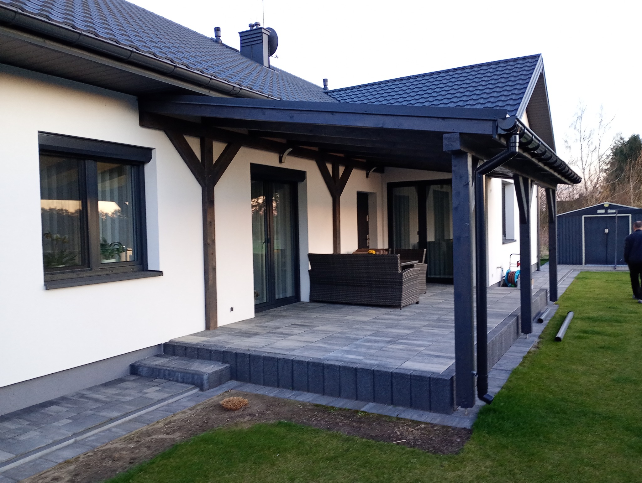 Zadaszony taras z ciemną konstrukcją drewnianą i szarą kostką brukową. Widoczny fragment dachu z rynnami i budynek gospodarczy w tle.