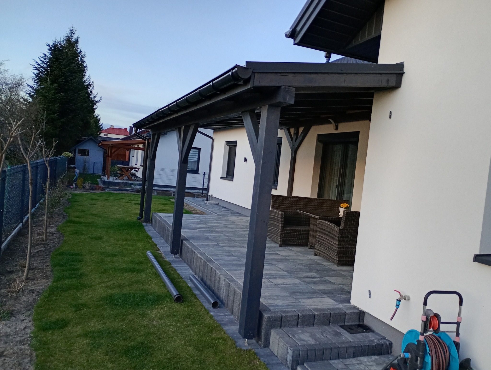 Zadaszony taras z ciemnymi elementami drewnianymi i szarą kostką brukową. Widoczne rury spustowe, meble ogrodowe i trawnik. Ujęcie z boku, w tle zabudowa jednorodzinna.