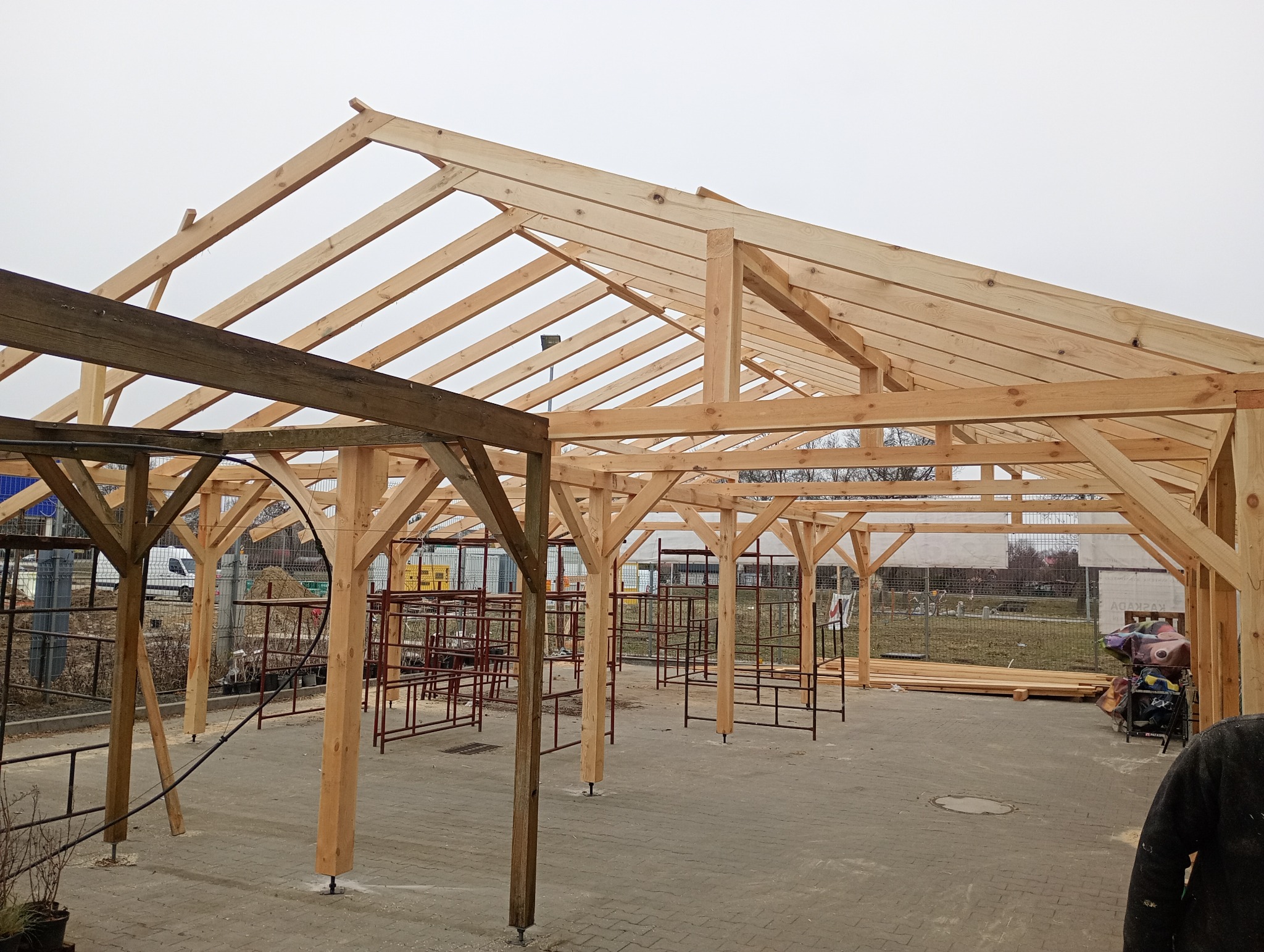 Surowa drewniana konstrukcja dachu z widoczną więźbą, słupami i belkami, na tle szarego nieba. Elementy metalowe w tle. Widok z poziomu gruntu.