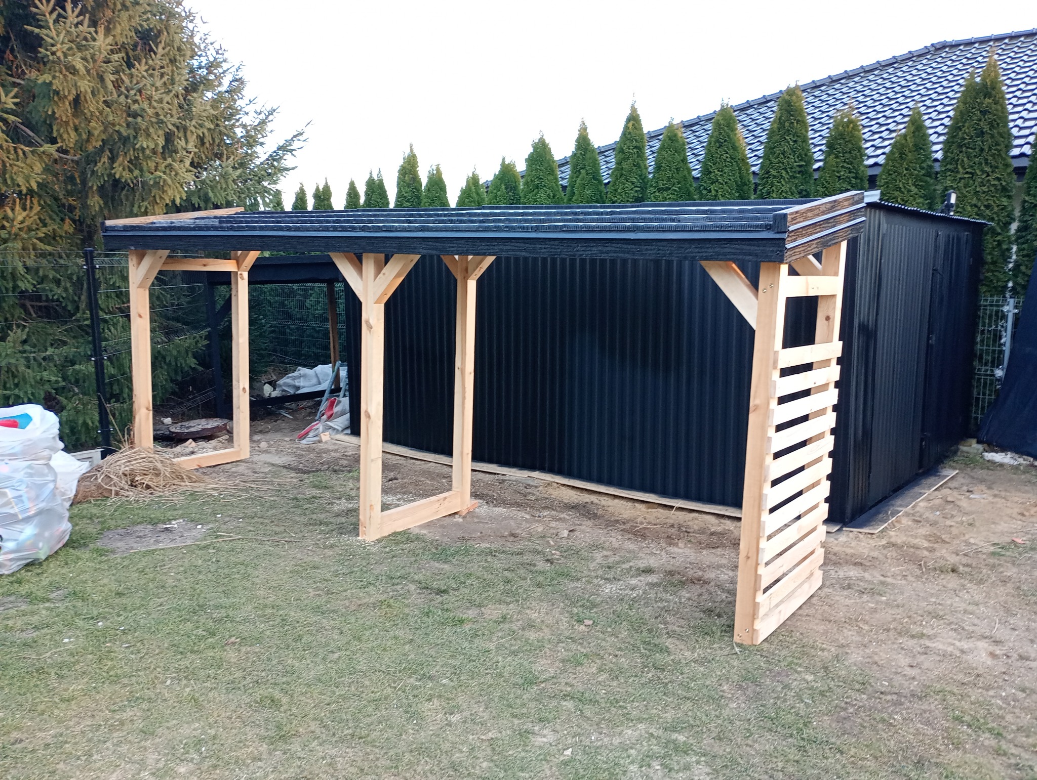 Konstrukcja zadaszenia z czarną blachą trapezową i drewnianymi słupami, widok z zewnątrz na tle zieleni i budynku z dachem dwuspadowym.