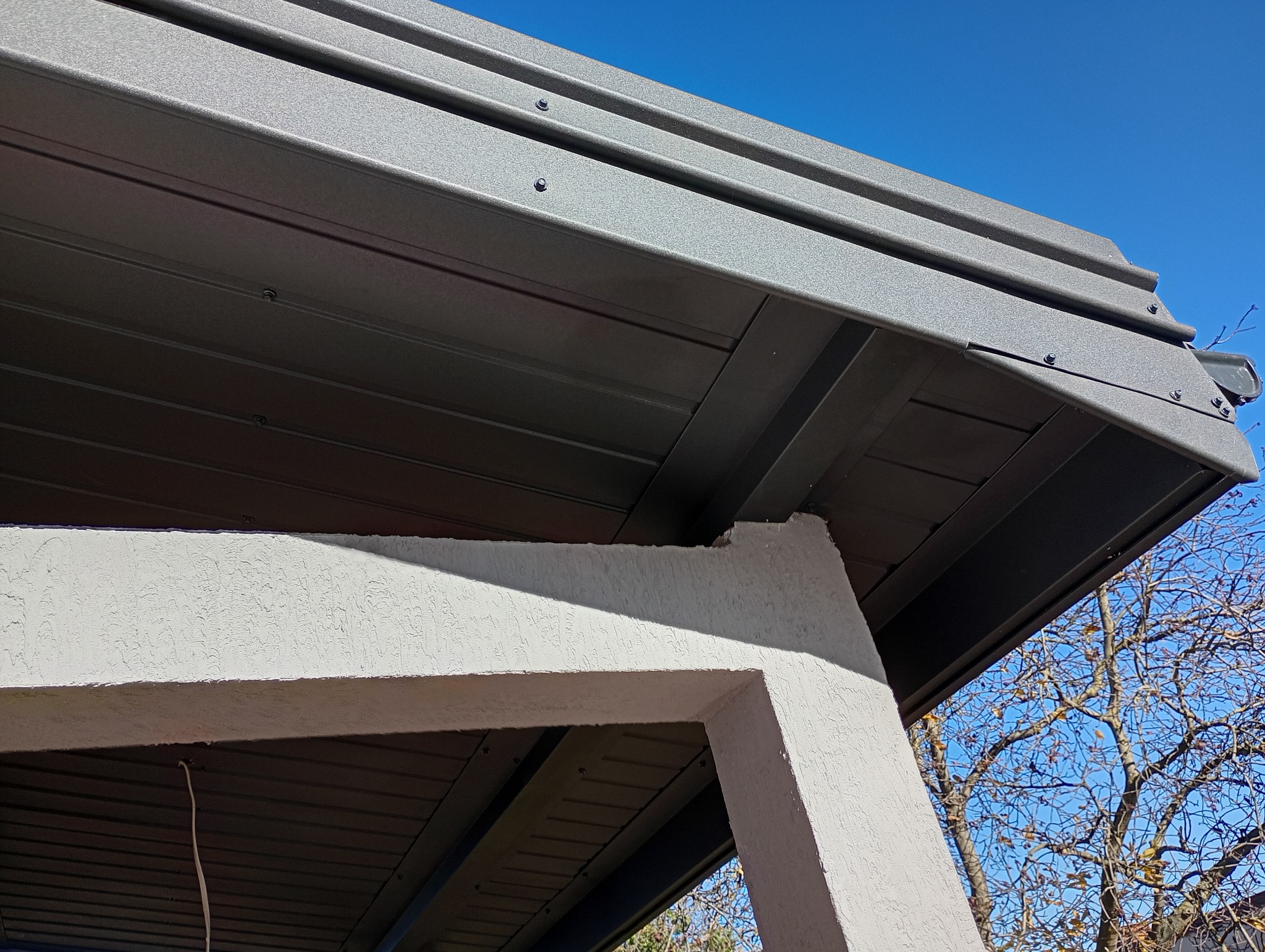 Szara, metalowa konstrukcja z widocznymi śrubami, prawdopodobnie zadaszenie tarasu, wsparta na białym, teksturowanym filarze. W tle błękitne niebo i gałęzie drzew.