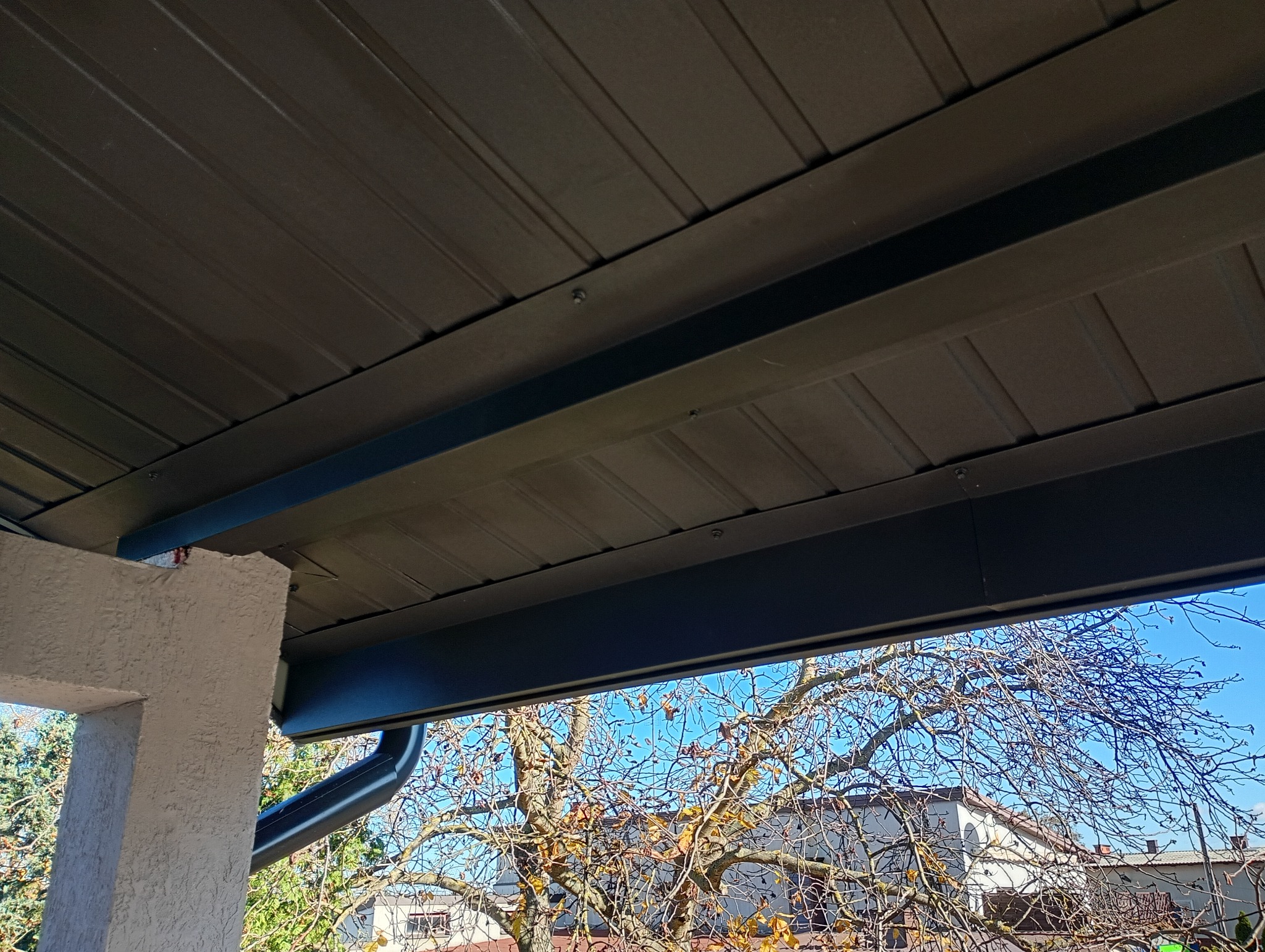 Metalowe pokrycie dachu z widoczną konstrukcją wsporczą i rynną, z perspektywy od dołu, z fragmentem ściany budynku i drzewem w tle.