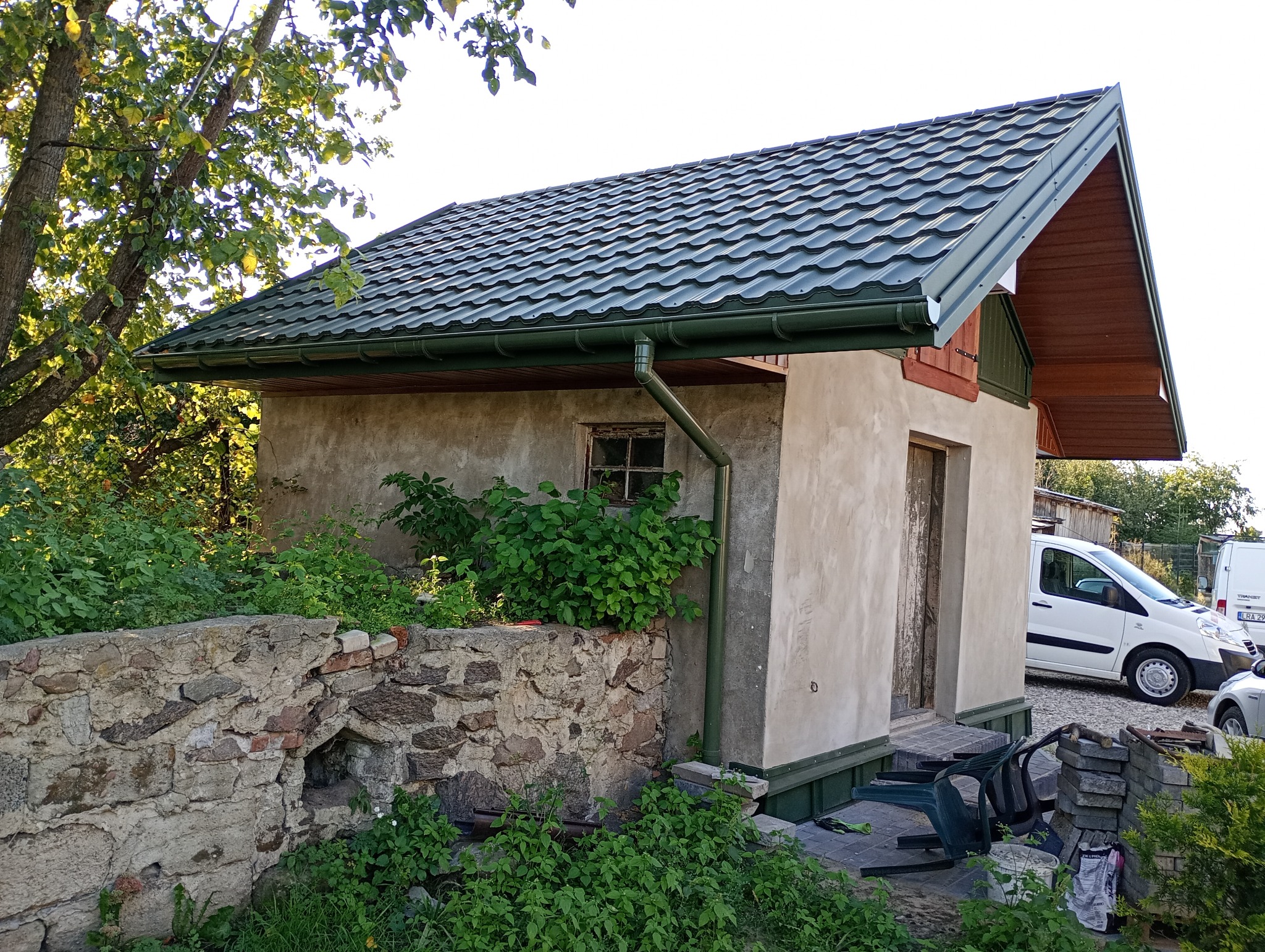 Mały budynek z nowym, zielonym dachem z blachy trapezowej i rynnami w tym samym kolorze. Widoczny fragment kamiennego murku i zarośla.