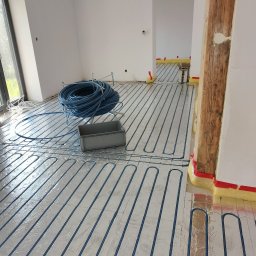 MIM INSTALACJE - Niebieskie rury ogrzewania podłogowego ułożone na srebrnej folii izolacyjnej w jasnym pomieszczeniu z drewnianym słupem. Część rur zwinięta w krąg obok szarego pojemnika.