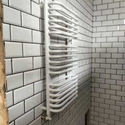 MIM INSTALACJE - Biały, drabinkowy grzejnik łazienkowy na ścianie z białych, prostokątnych płytek. Widoczne przyłącze. Fragment ściany bez płytek w trakcie prac wykończeniowych.